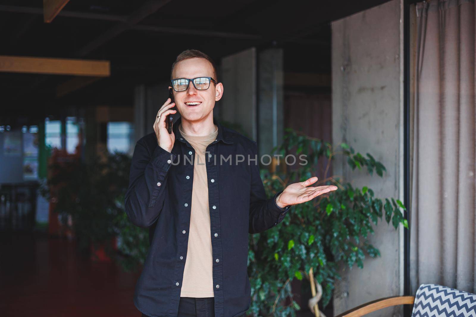 Half-length portrait of happy man in optical glasses holding modern mobile phone and smiling at camera, cheerful blogger with digital smartphone posing in coworking space