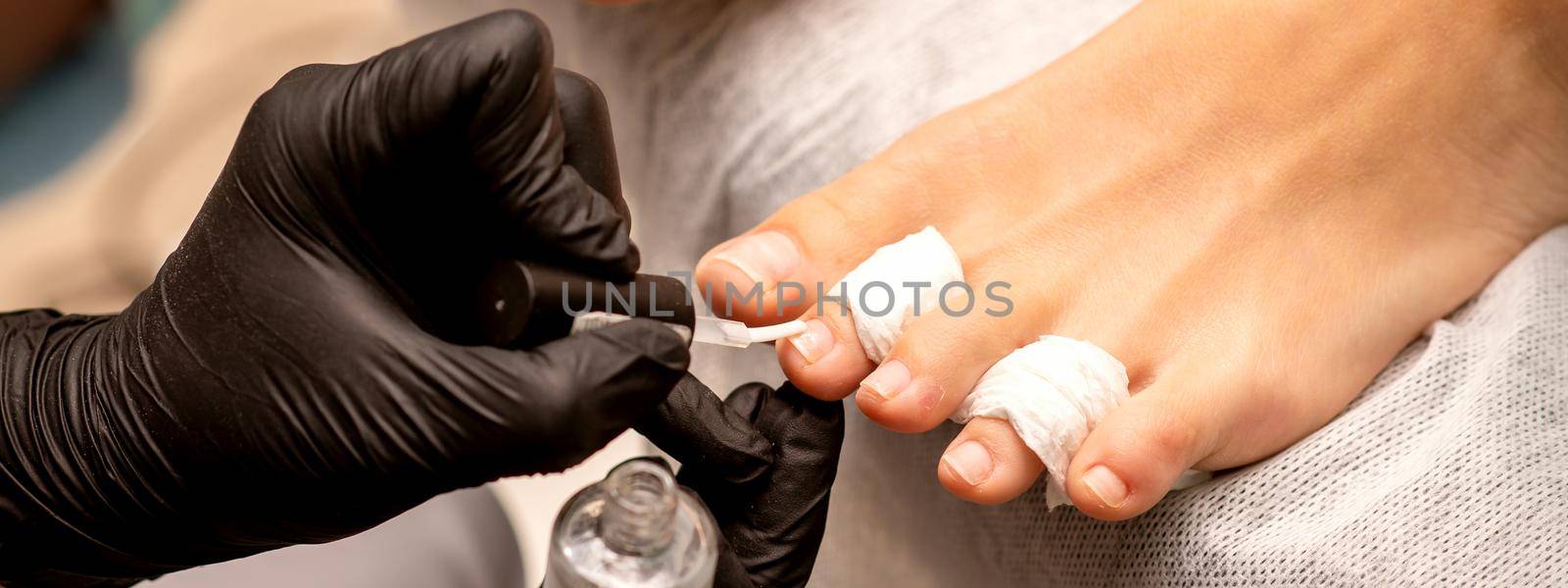 Manicure master painting nails on a female foot with transparent nail polish in a beauty salon, close up. by okskukuruza