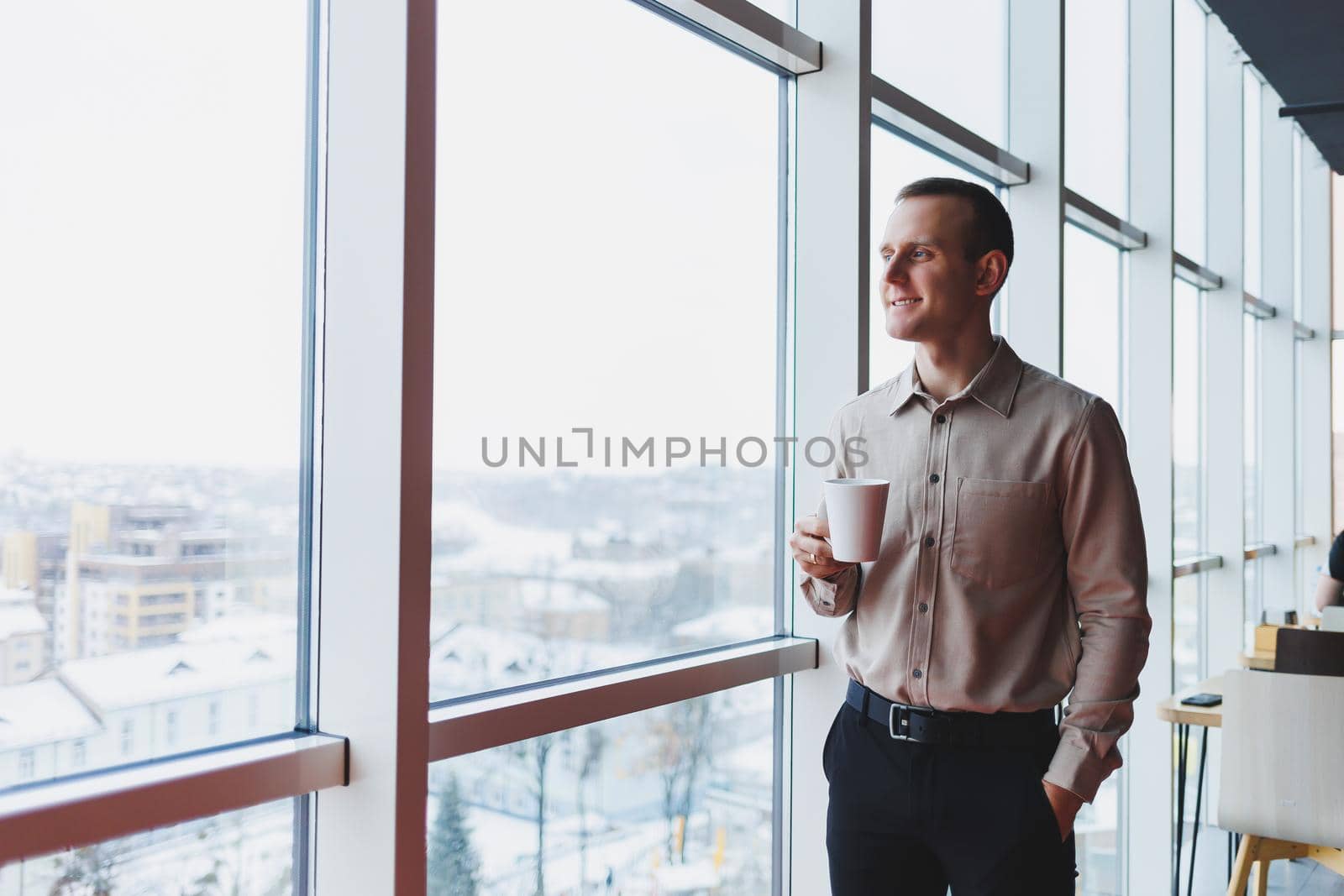 Men in shirt and trousers looking towards window with coffee in hand, in modern workspace