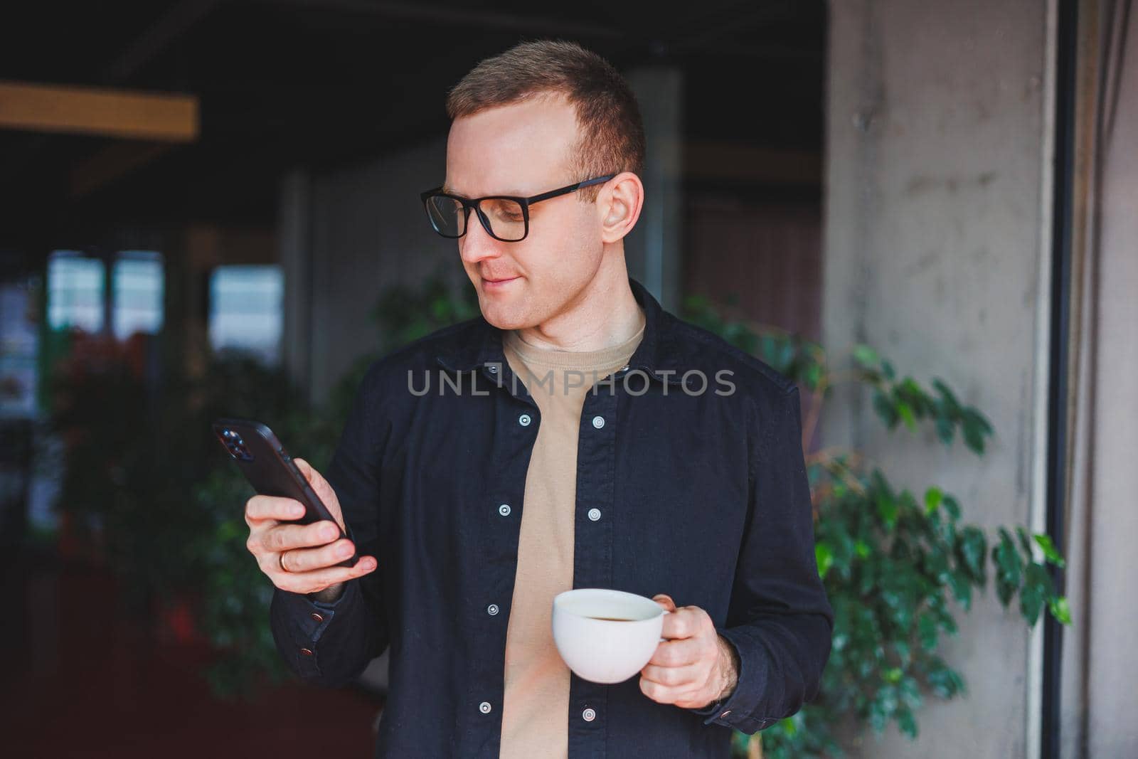 Portrait of a happy successful man in optical glasses holding a modern mobile phone and coffee in his hands, a cheerful blogger with a digital smartphone posing in a cafe