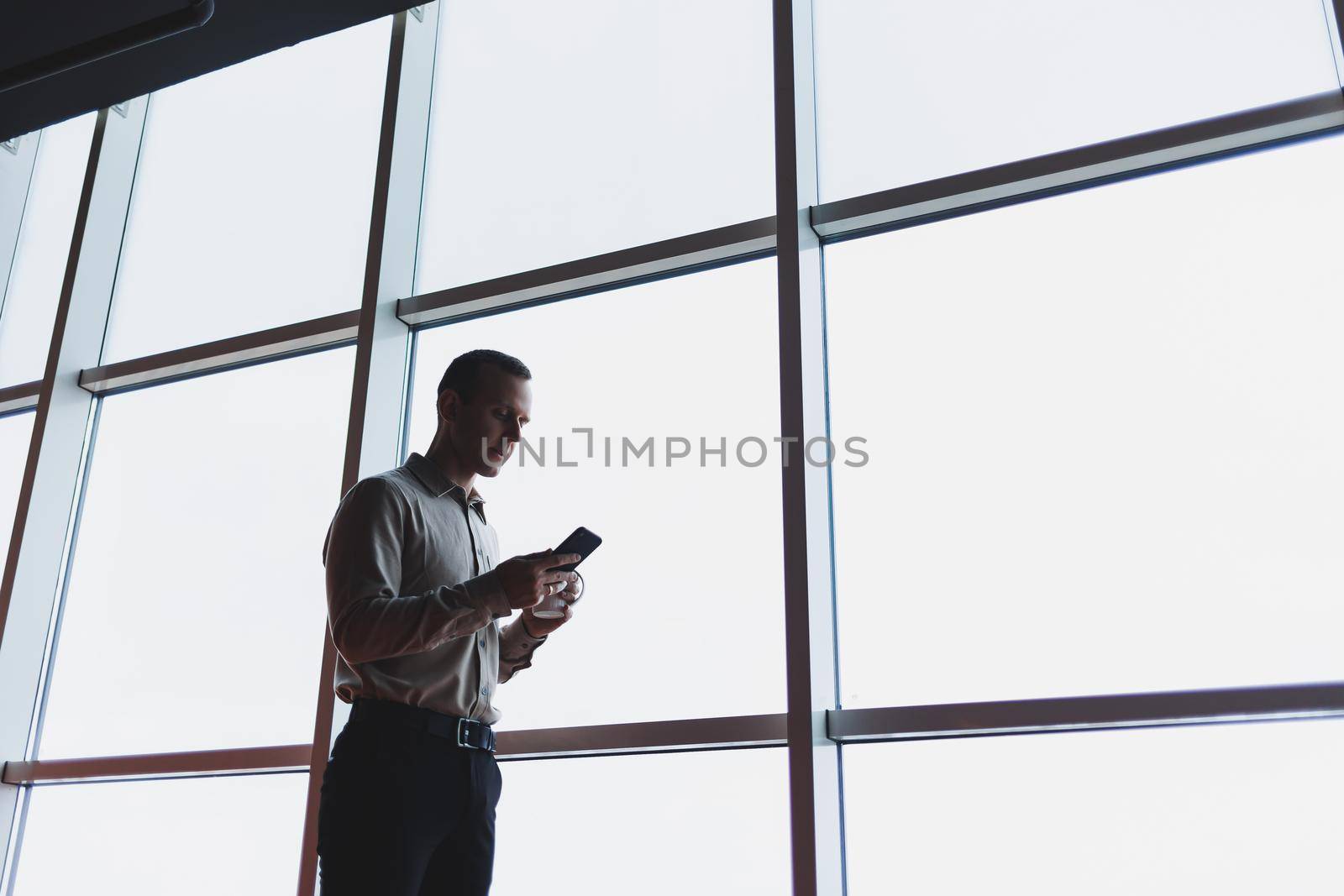 A young executive is holding a phone while standing in an office interior and looking out a large window overlooking the city. Male manager with a phone is thinking about a new business project