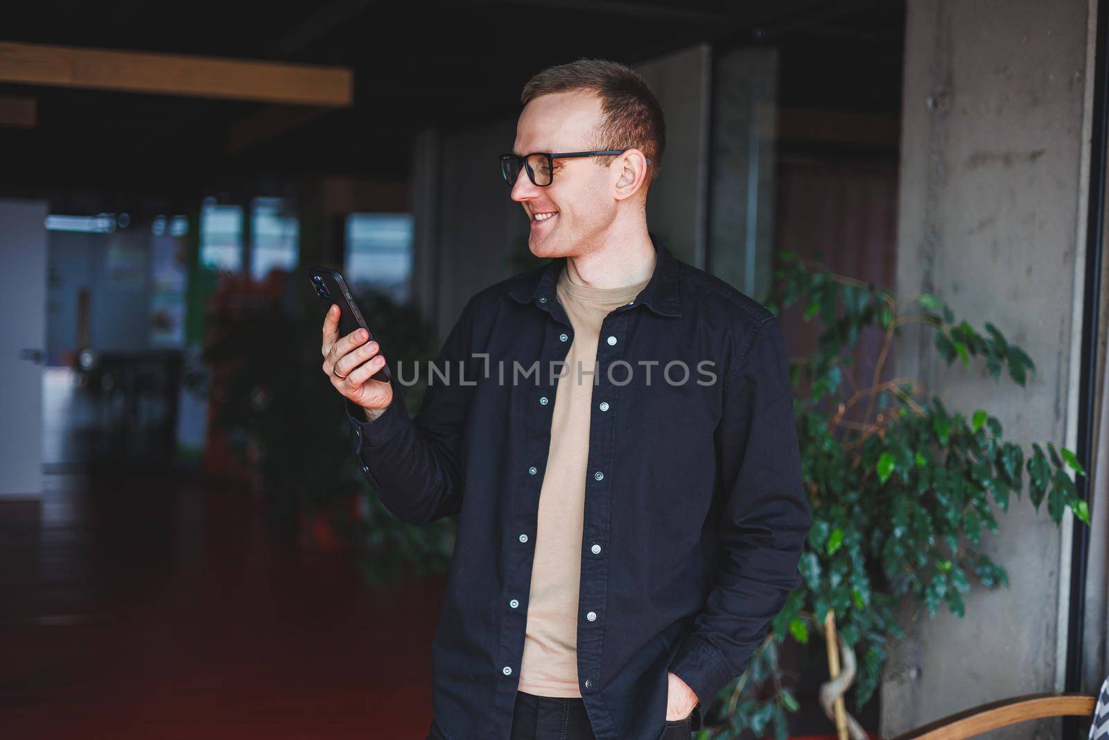 Half-length portrait of happy man in optical glasses holding modern mobile phone and smiling at camera, cheerful blogger with digital smartphone posing in coworking space