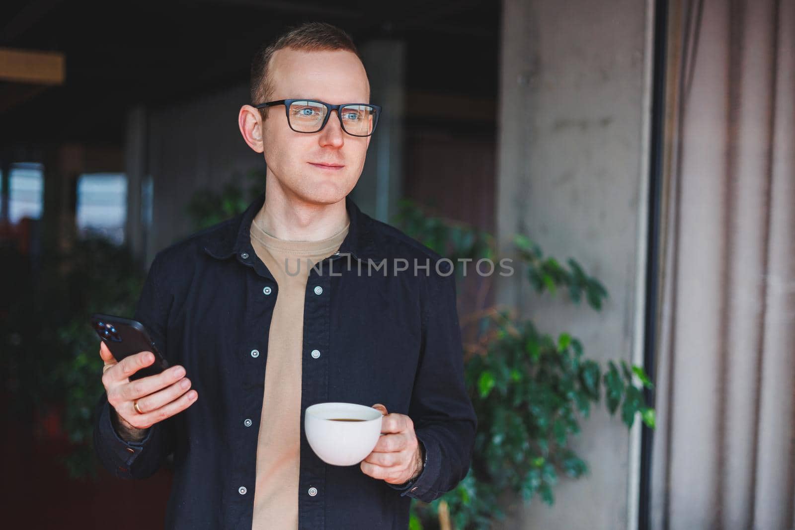 Portrait of a happy successful man in optical glasses holding a modern mobile phone and coffee in his hands, a cheerful blogger with a digital smartphone posing in a cafe