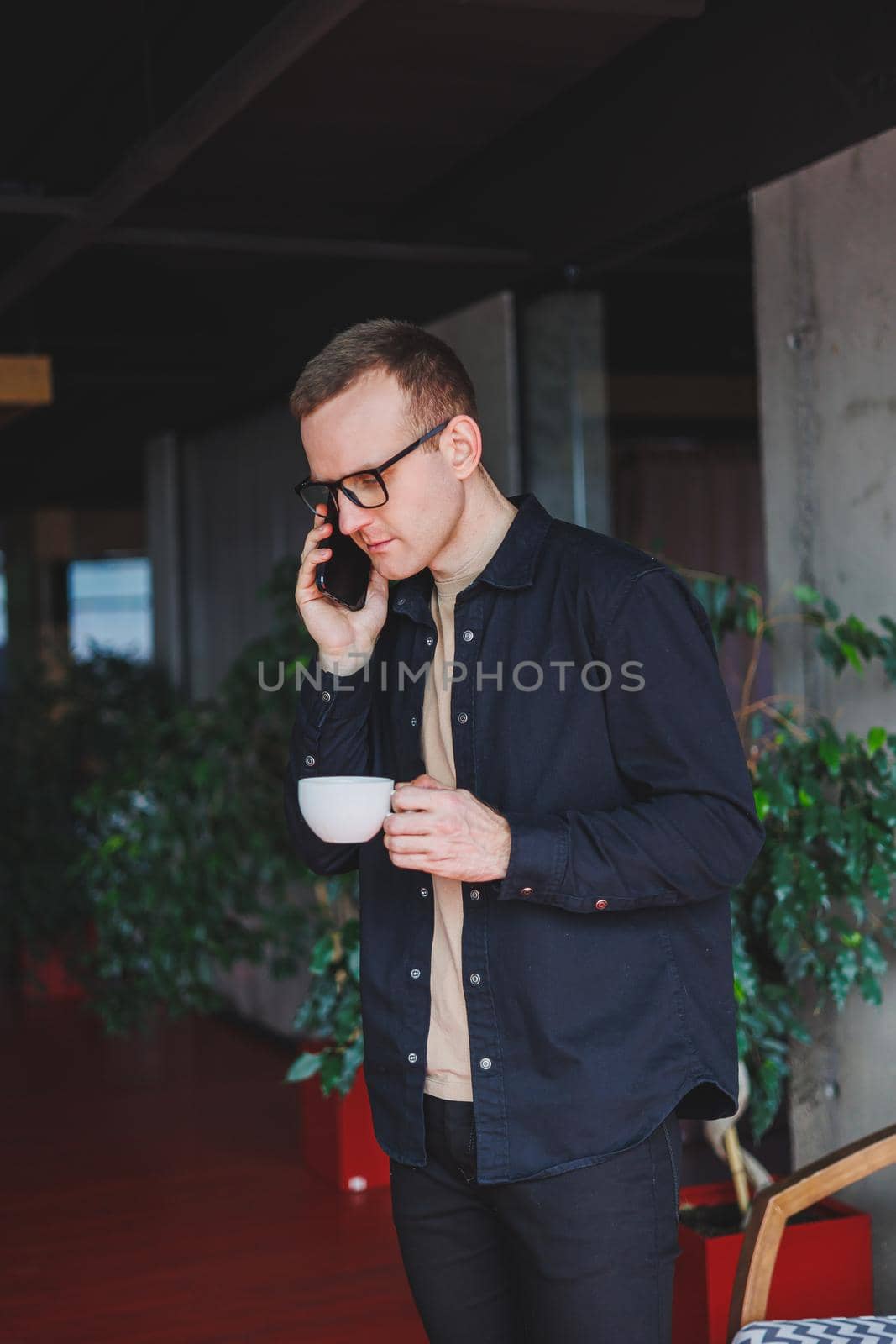 Portrait of a happy successful man in optical glasses holding a modern mobile phone and coffee in his hands, a cheerful blogger with a digital smartphone posing in a cafe