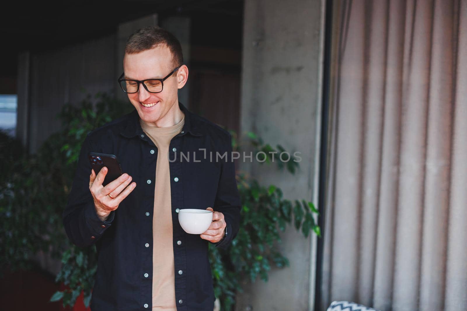 Portrait of a happy successful man in optical glasses holding a modern mobile phone and coffee in his hands, a cheerful blogger with a digital smartphone posing in a cafe