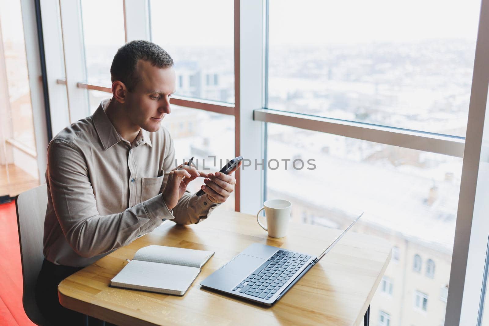 Successful young man is sitting at a table in a cafe-restaurant indoors, working and studying on a laptop computer,holding a mobile phone with a blank screen. Freelancer mobile office business concept