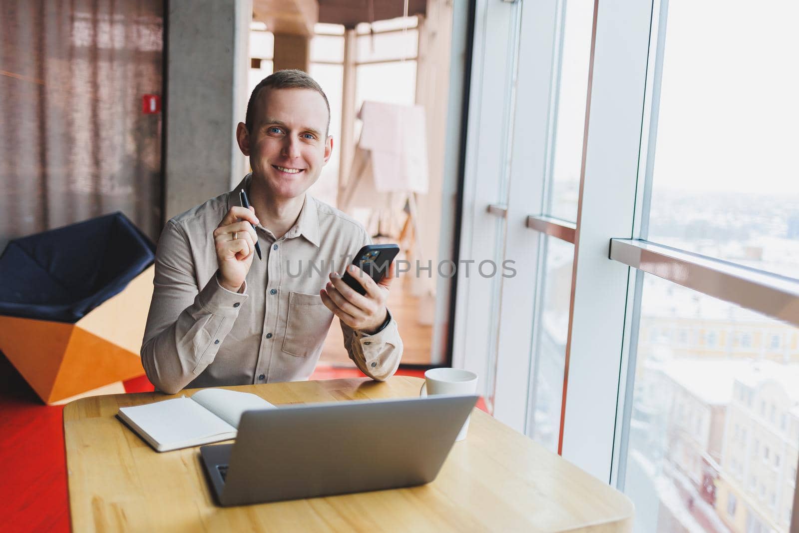 Successful young man is sitting at a table in a cafe-restaurant indoors, working and studying on a laptop computer,holding a mobile phone with a blank screen. Freelancer mobile office business concept