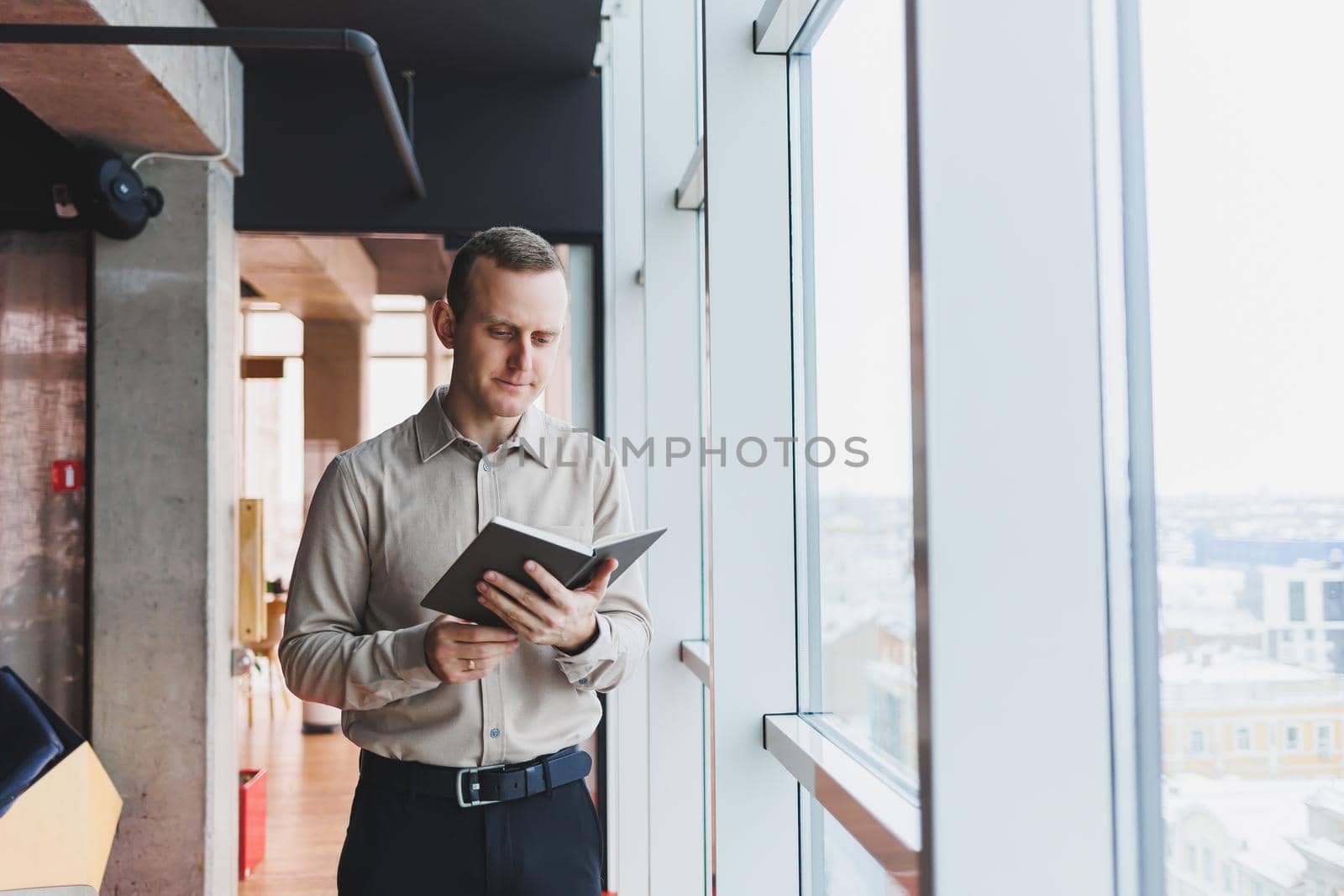 A young businessman stands at a panoramic window in a skyscraper and makes notes in his notebook with a pen. A man in a shirt and trousers of European appearance