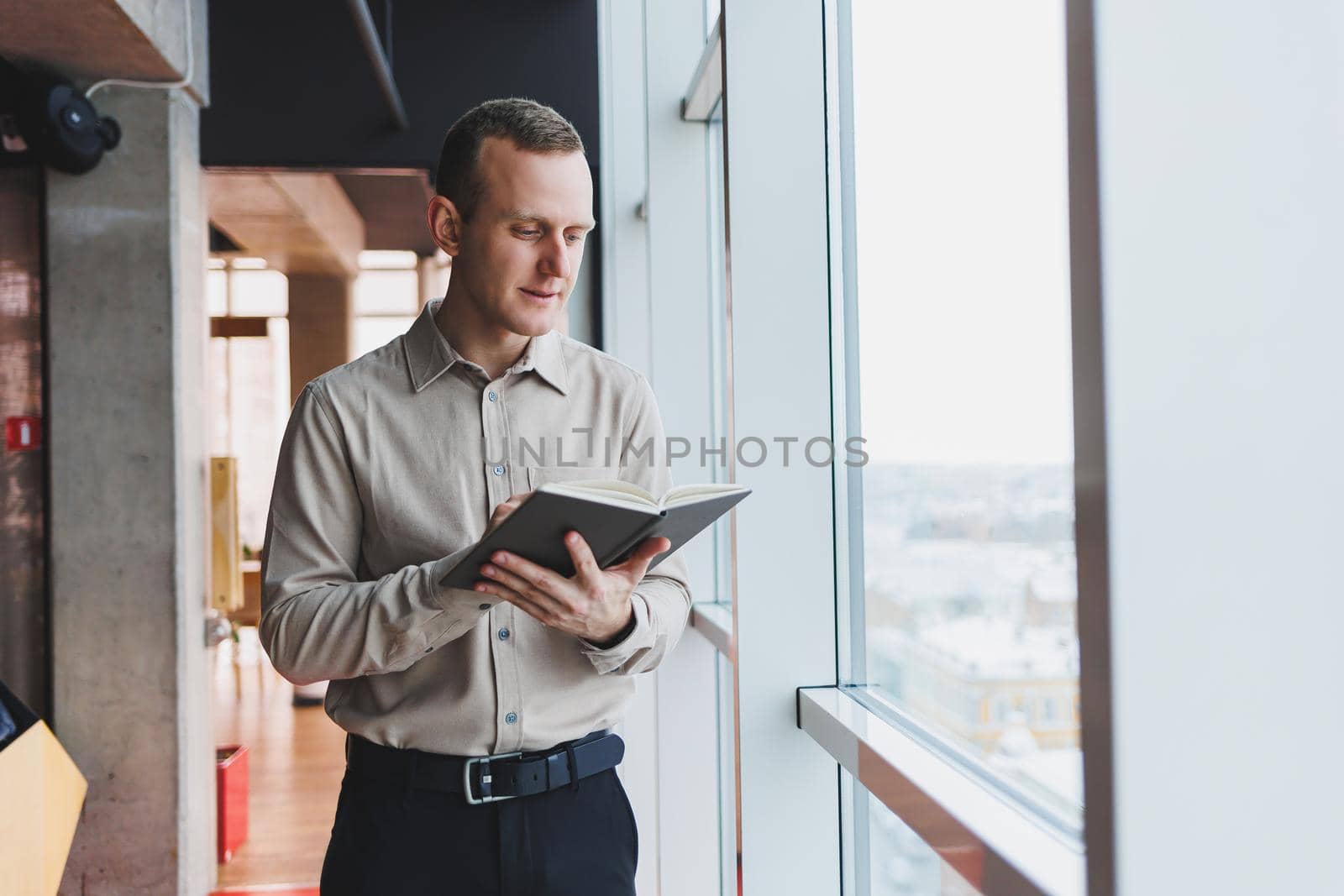 A young businessman stands at a panoramic window in a skyscraper and makes notes in his notebook with a pen. A man in a shirt and trousers of European appearance