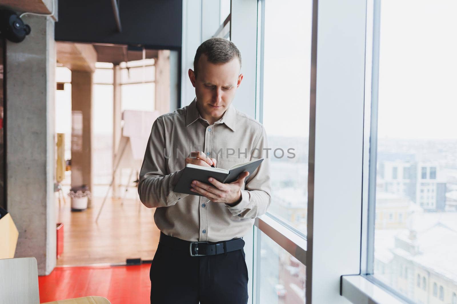 A young businessman stands at a panoramic window in a skyscraper and makes notes in his notebook with a pen. A man in a shirt and trousers of European appearance