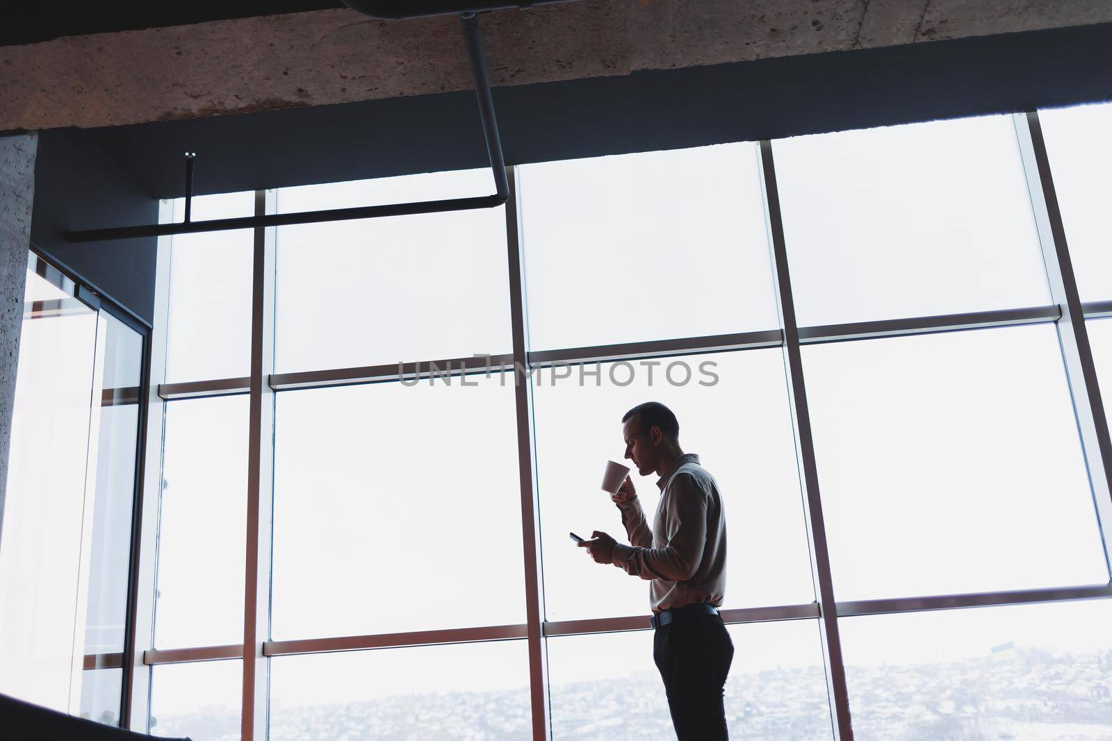 A young executive is holding a phone while standing in an office interior and looking out a large window overlooking the city. Male manager with a phone is thinking about a new business project