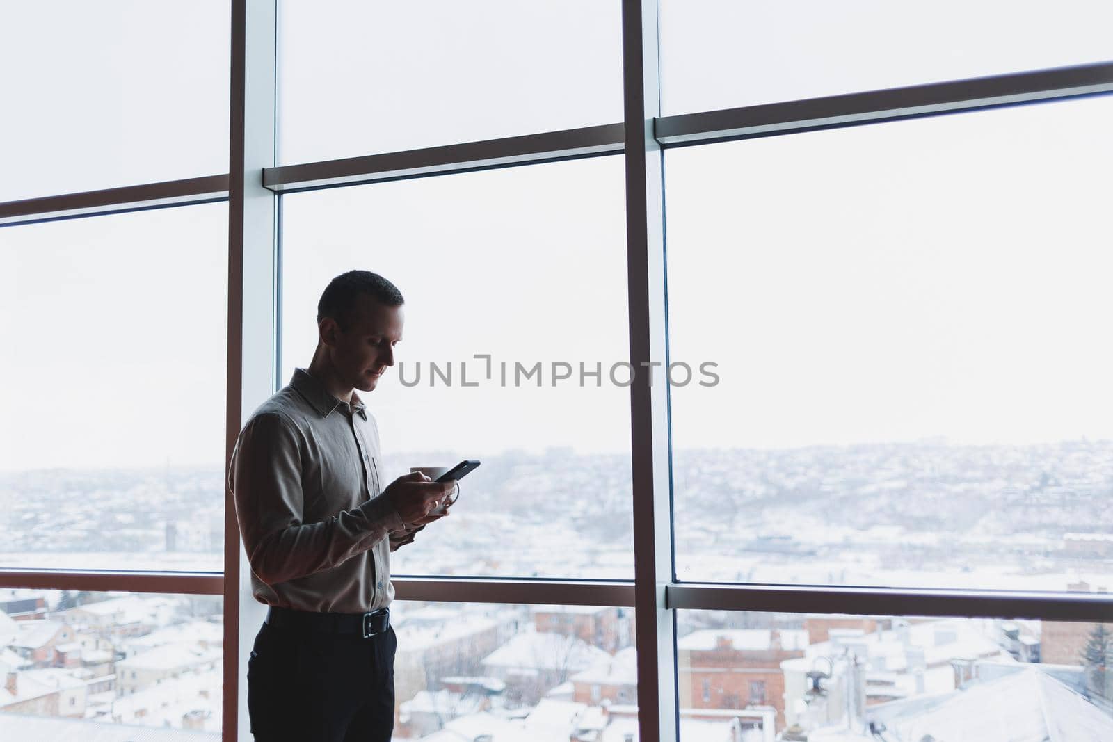 A young executive is holding a phone while standing in an office interior and looking out a large window overlooking the city. Male manager with a phone is thinking about a new business project