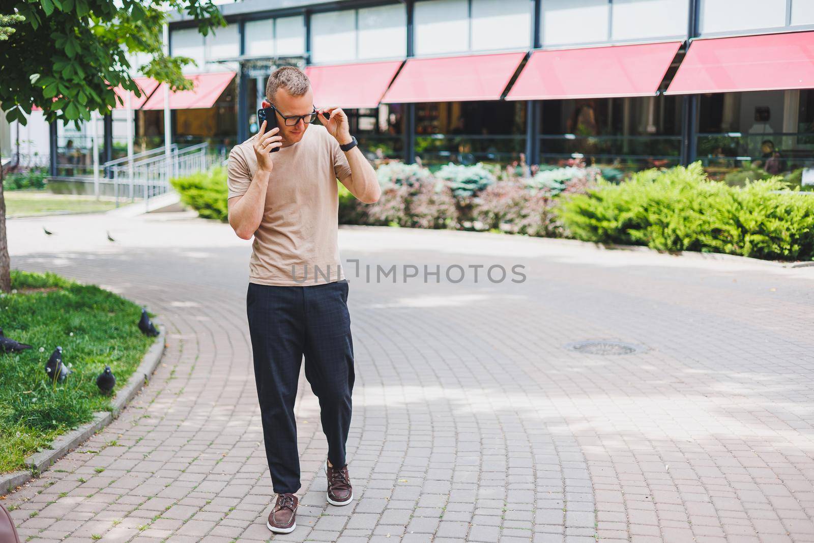 Portrait of handsome mature businessman holding messenger bag, wearing stylish eyeglasses looking at camera. Smiling middle aged man using smartphone, standing on the street. Mobile banking concept