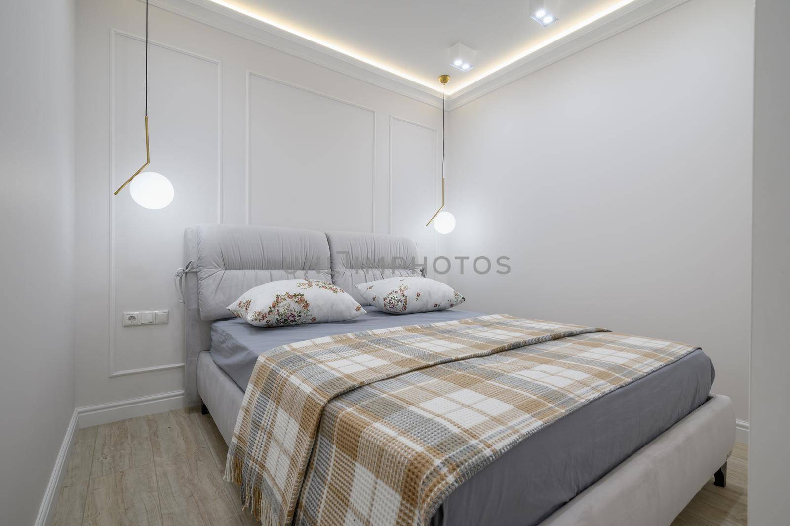Double bed with a plaid blanket and pillows on it in a cozy bedroom with white walls and wood floors and a light fixtures