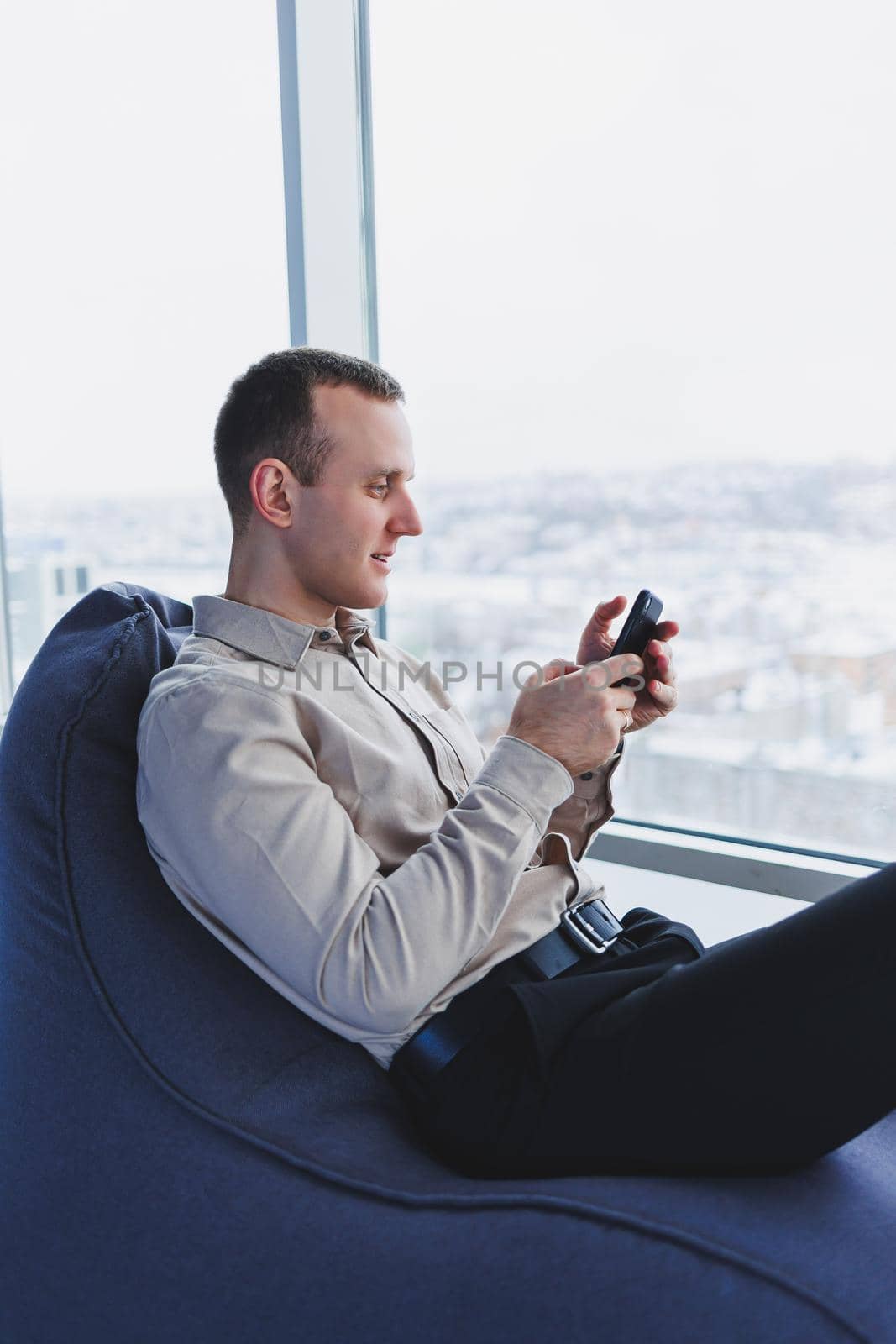 Successful male businessman sitting in a modern workspace and checking notifications on his mobile phone, he is wearing a shirt and trousers. Remote work.