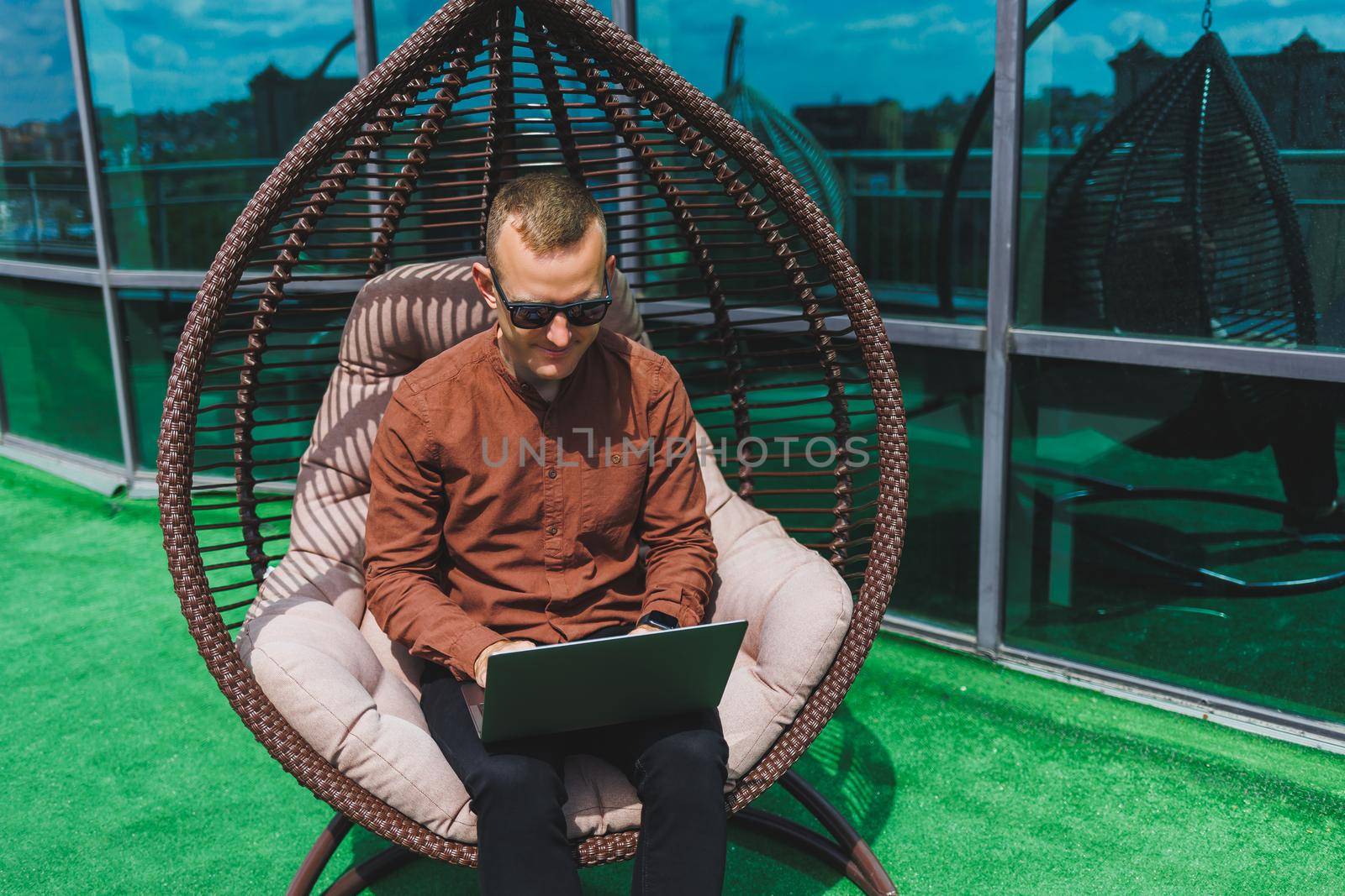 A businessman in formal wear sits on an armchair and looks at a laptop connected to a 4g wireless network on the terrace of a modern office while working remotely