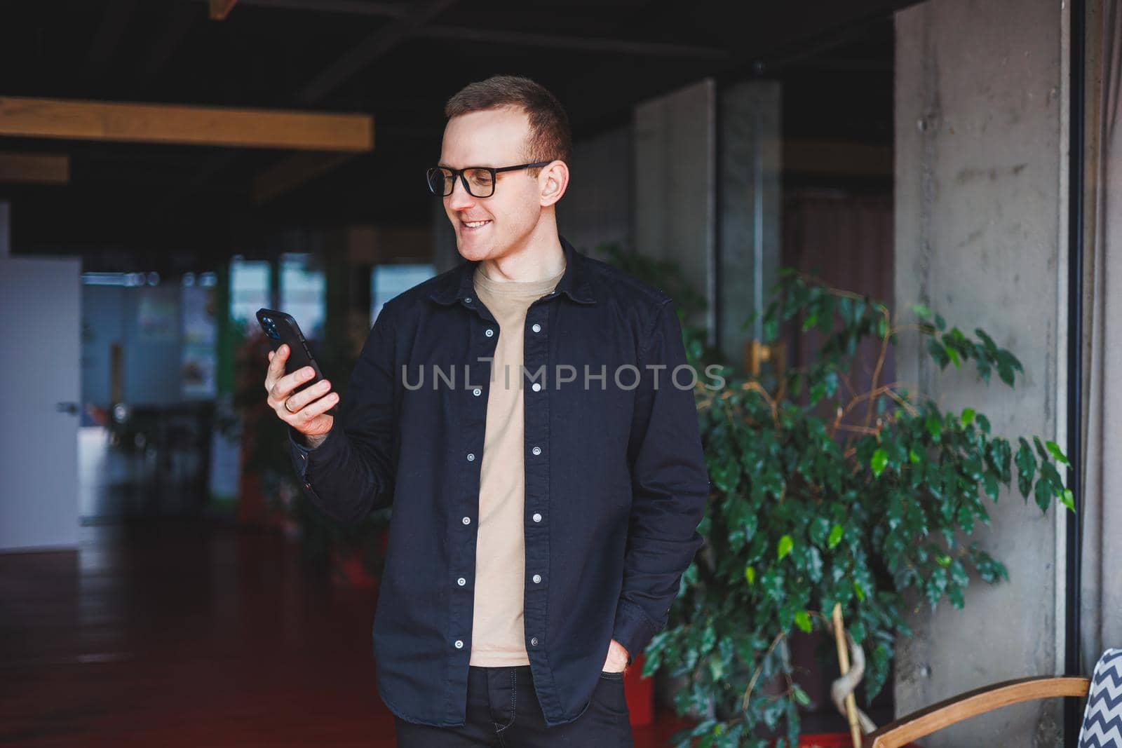 Half-length portrait of happy man in optical glasses holding modern mobile phone and smiling at camera, cheerful blogger with digital smartphone posing in coworking space