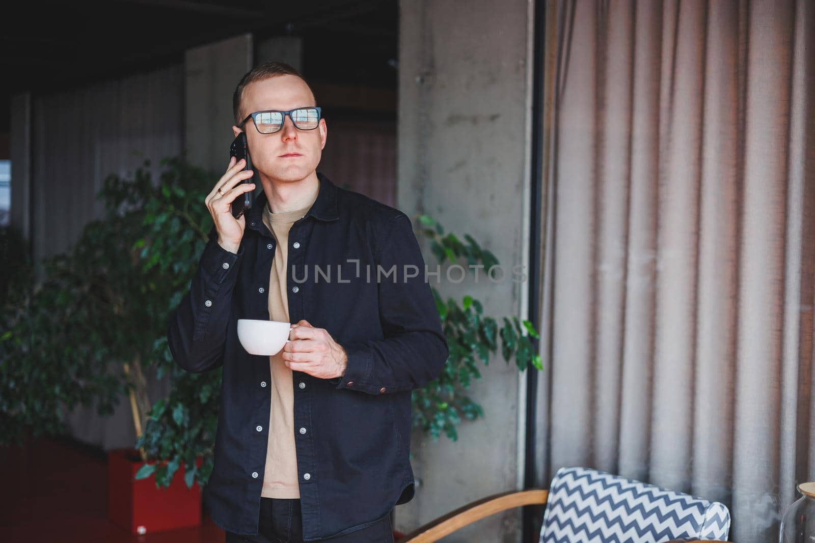 Portrait of a happy successful man in optical glasses holding a modern mobile phone and coffee in his hands, a cheerful blogger with a digital smartphone posing in a cafe