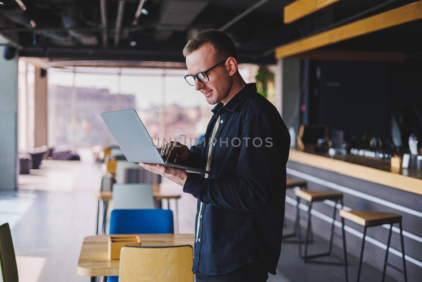 Male entrepreneur in black shirt and glasses standing in office with laptop in hand, successful corporate boss feeling good from rich lifestyle