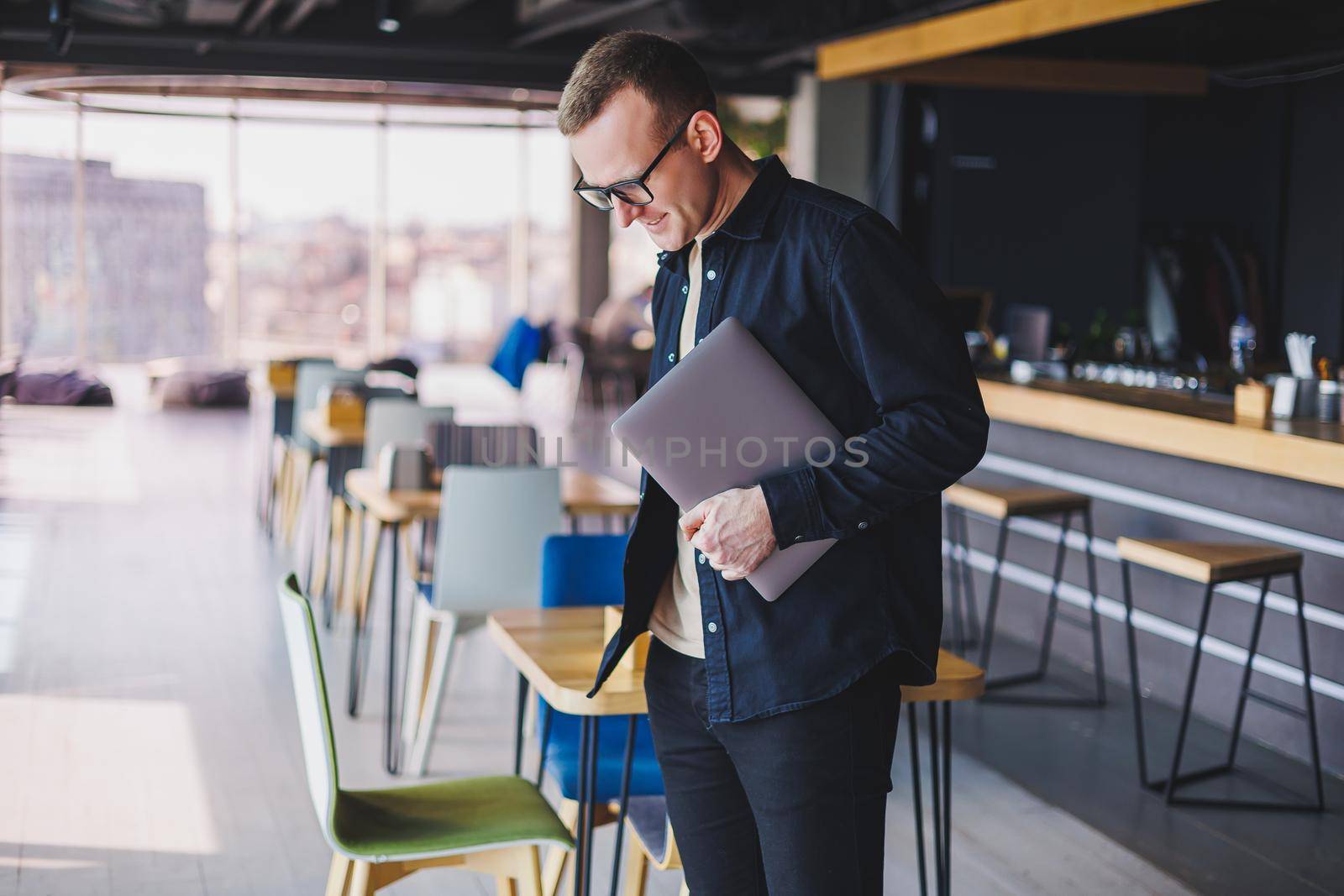 Male entrepreneur in black shirt and glasses standing in office with laptop in hand, successful corporate boss feeling good from rich lifestyle