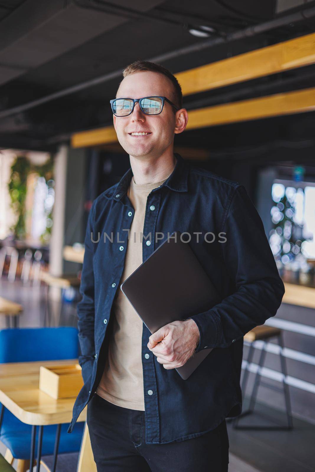 Male entrepreneur in black shirt and glasses standing in office with laptop in hand, successful corporate boss feeling good from rich lifestyle