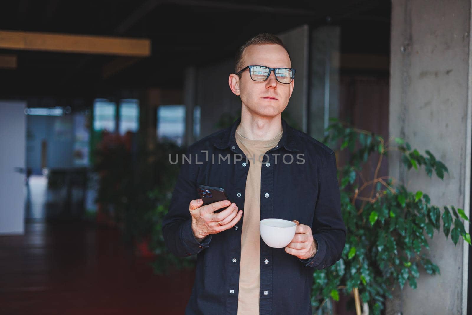Portrait of a happy successful man in optical glasses holding a modern mobile phone and coffee in his hands, a cheerful blogger with a digital smartphone posing in a cafe