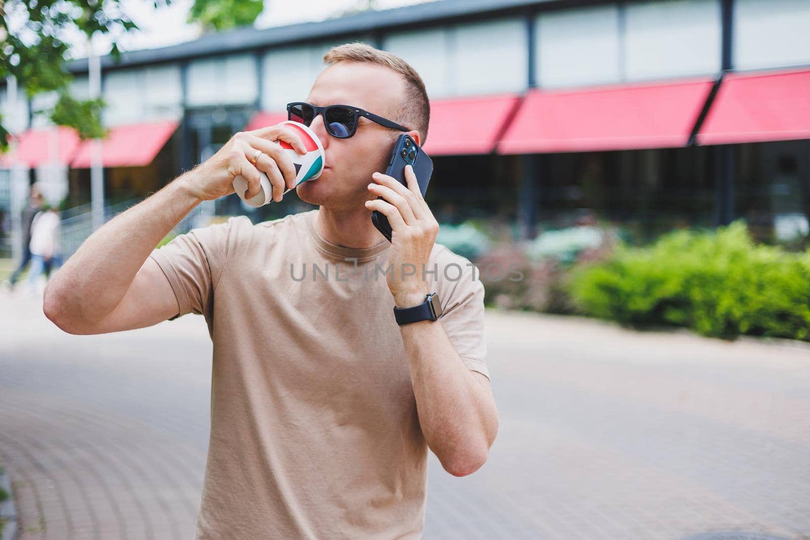 A bearded, smiling, stylish man in white shirt and sunglasses standing on the streets of the city and calling on mobile phone.