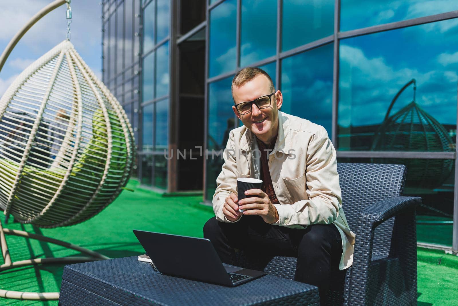 Confident young businessman, dressed in formal wear and stylish glasses, calls on a smartphone while sitting with coffee, on the terrace of a modern office building with a glass wall