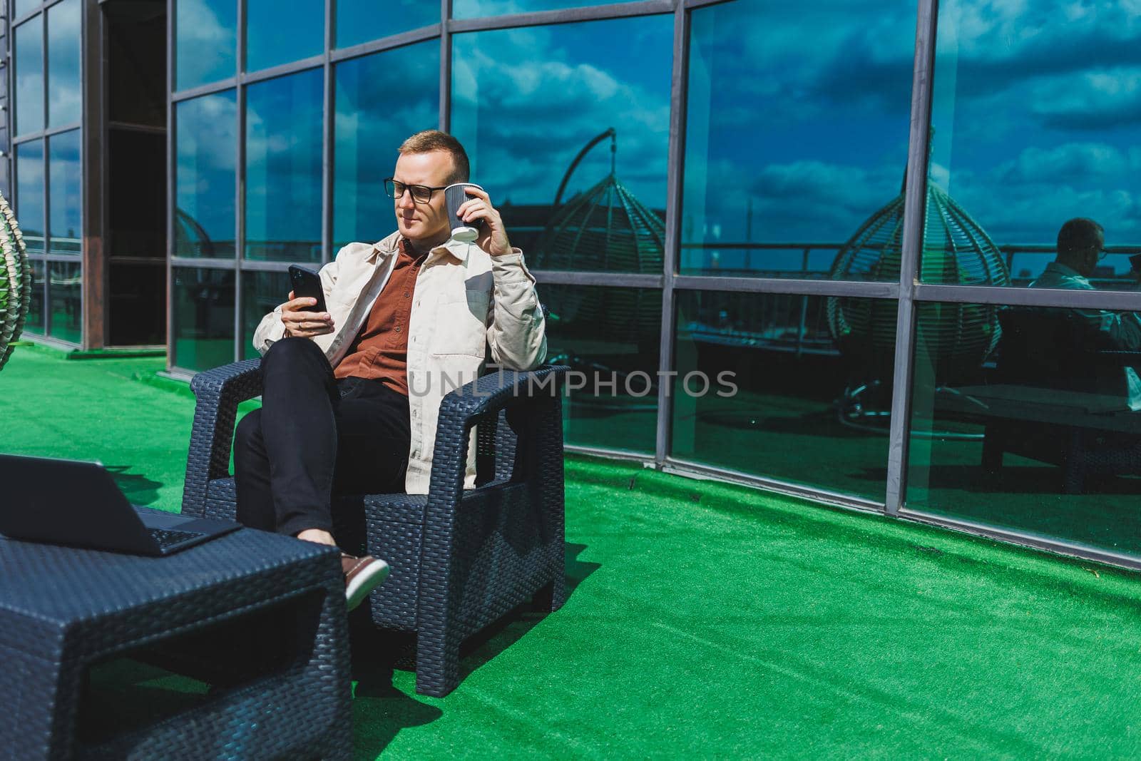 Confident young businessman, dressed in formal wear and stylish glasses, calls on a smartphone while sitting with coffee, on the terrace of a modern office building with a glass wall
