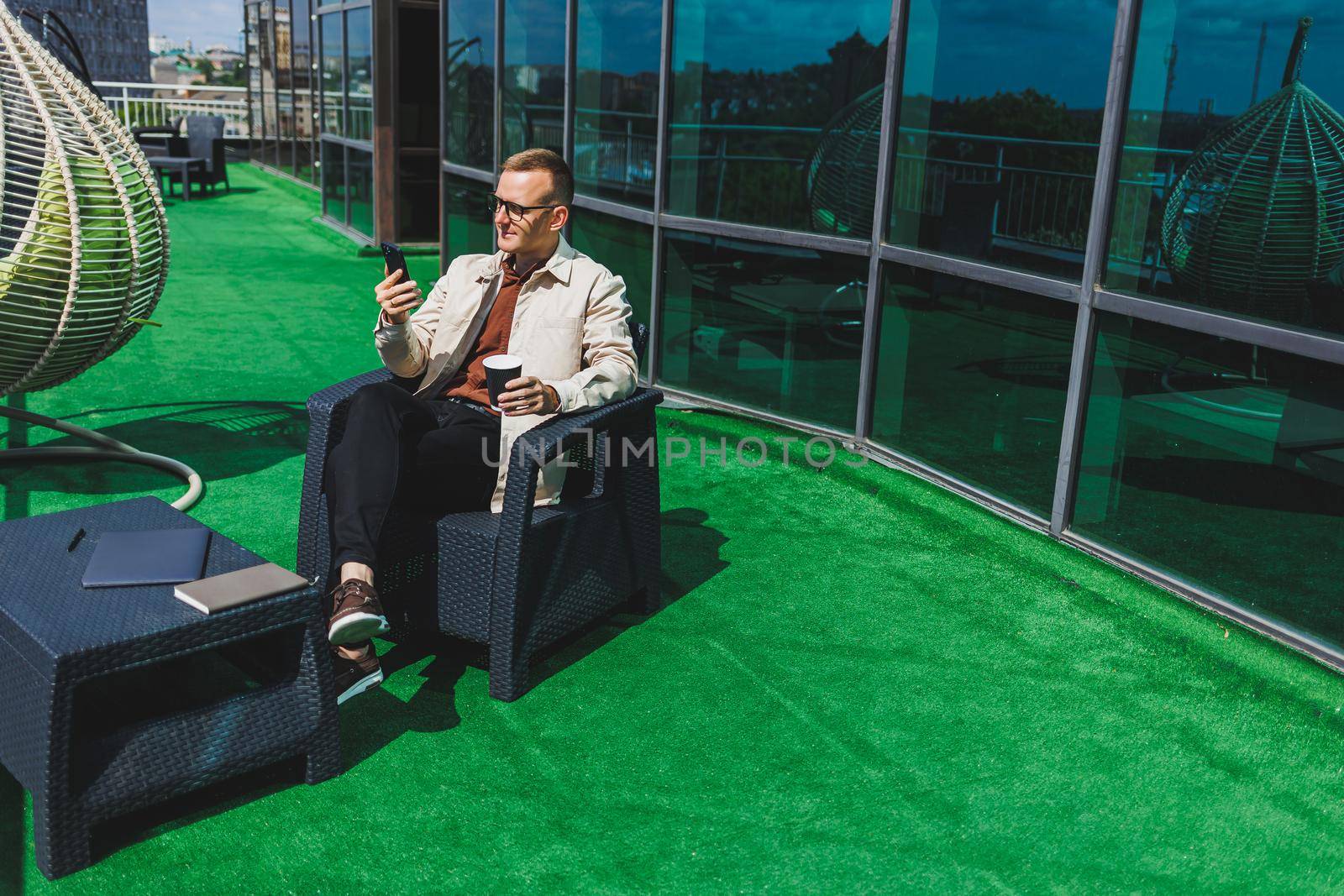 Confident young businessman, dressed in formal wear and stylish glasses, calls on a smartphone while sitting with coffee, on the terrace of a modern office building with a glass wall