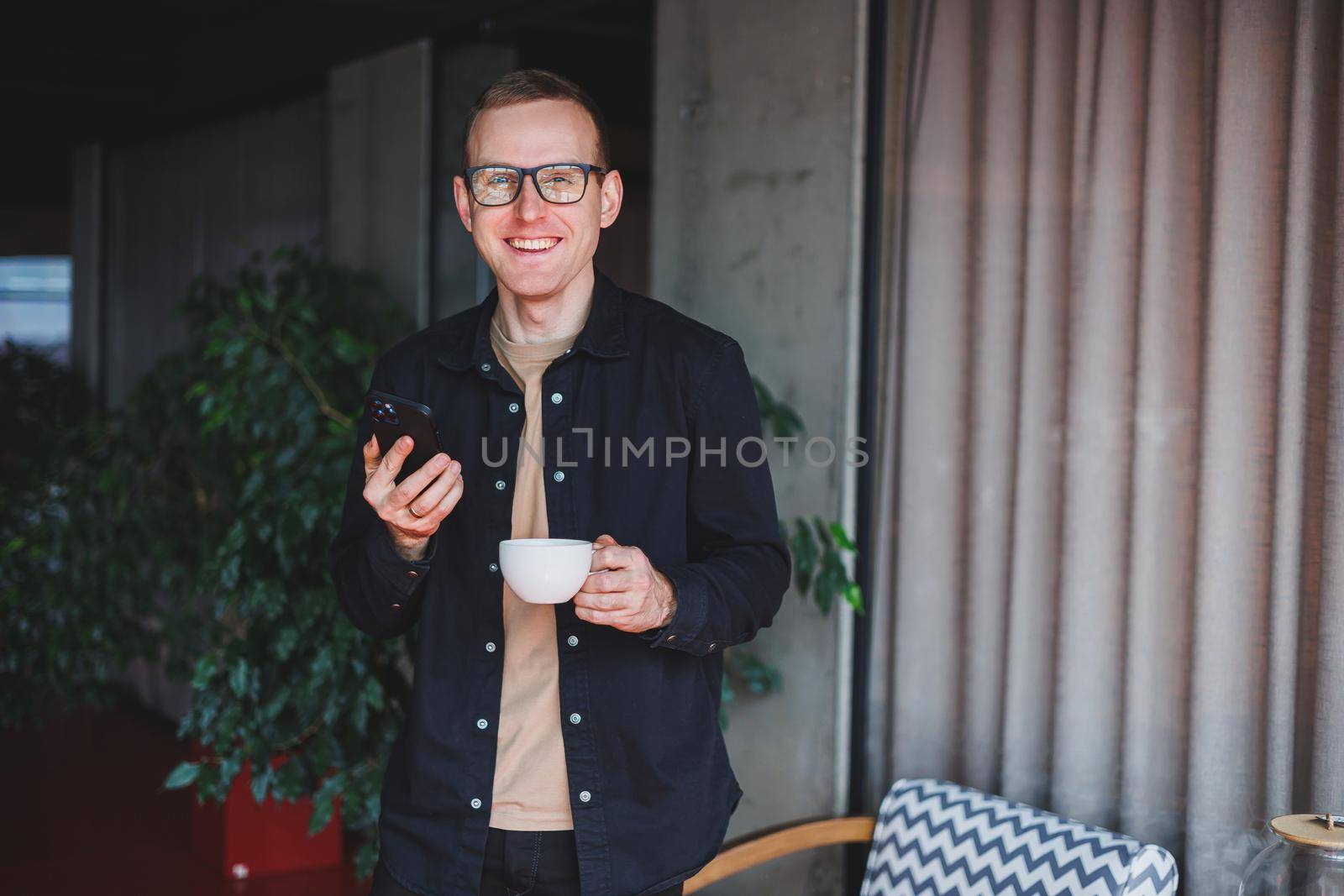 Portrait of a happy successful man in optical glasses holding a modern mobile phone and coffee in his hands, a cheerful blogger with a digital smartphone posing in a cafe
