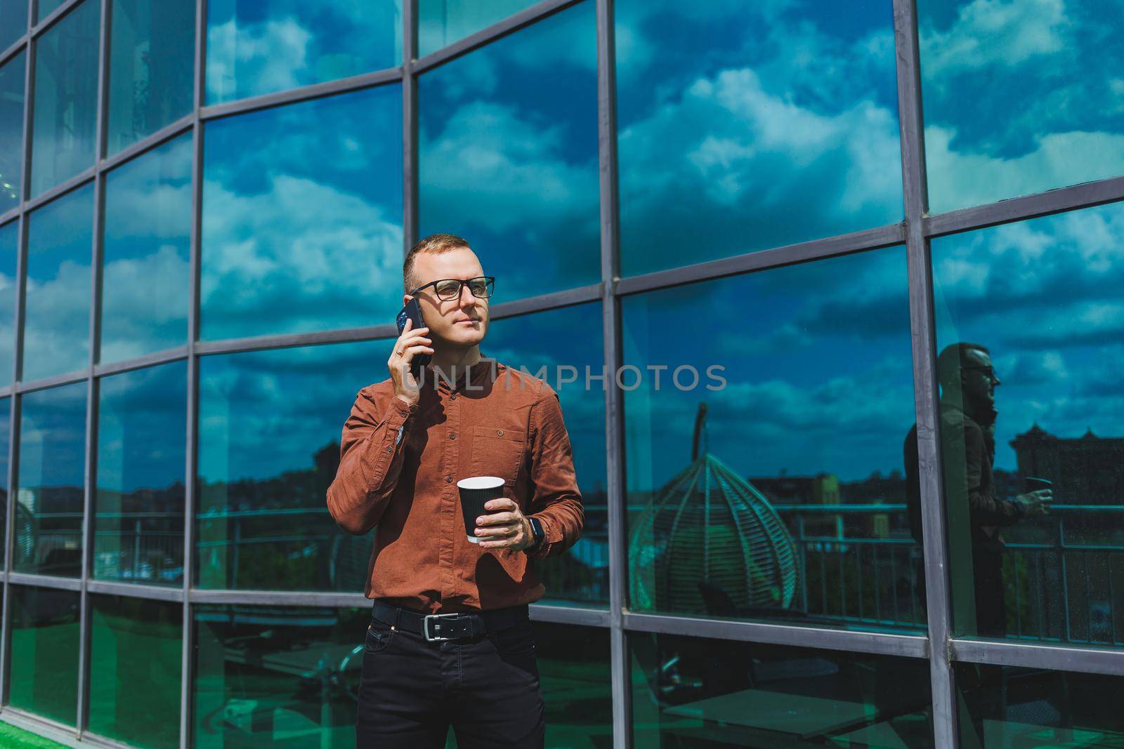 Attractive fashionable male businessman in a stylish shirt and trousers talking on the phone near the office building and drinking coffee, modern glass office