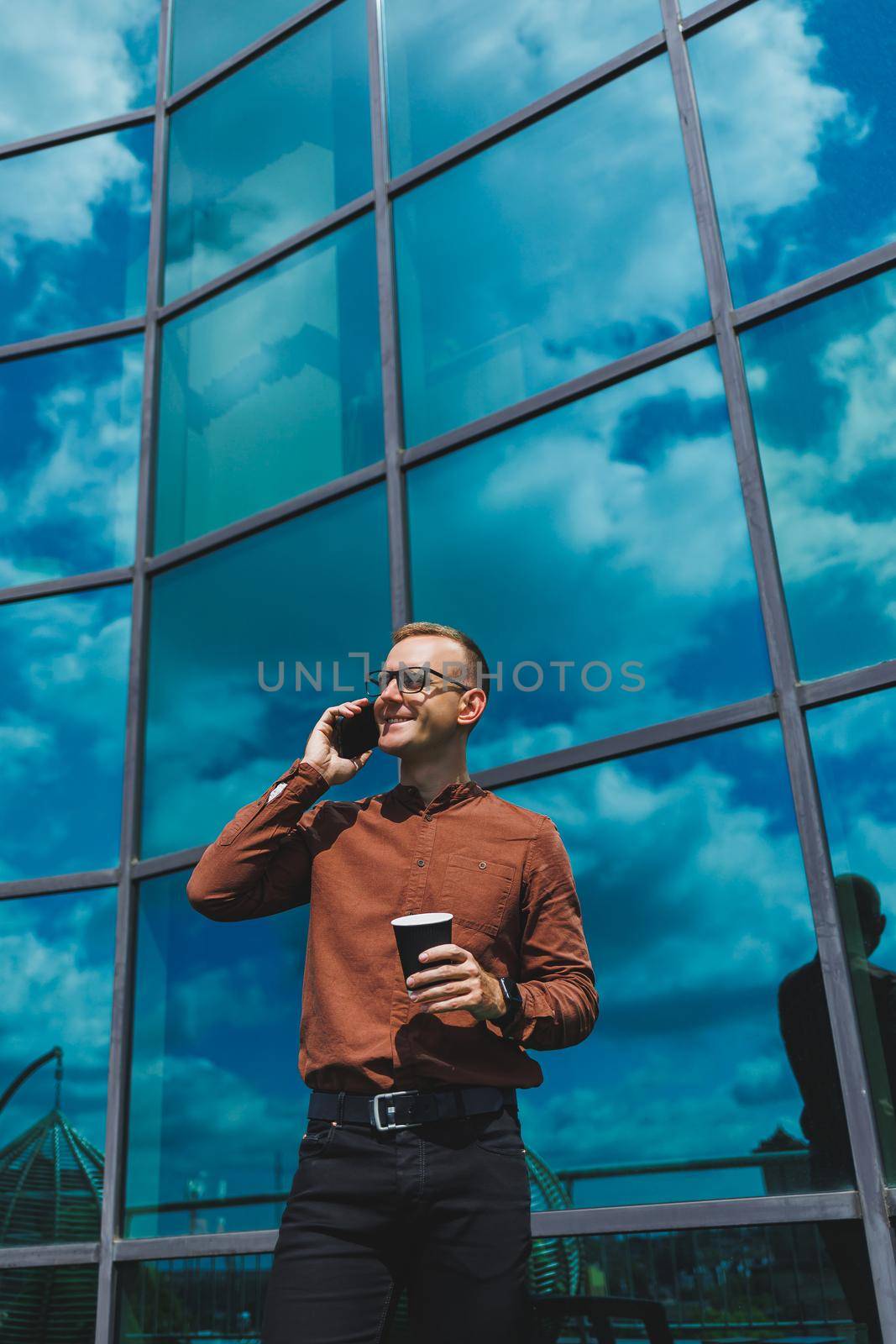 Attractive fashionable male businessman in a stylish shirt and trousers talking on the phone near the office building and drinking coffee, modern glass office