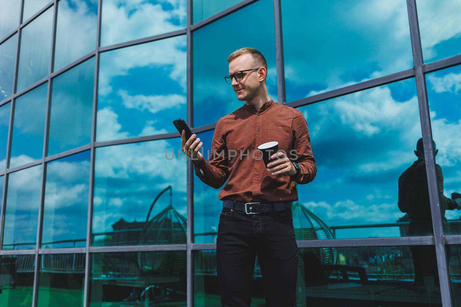 Attractive fashionable male businessman in a stylish shirt and trousers talking on the phone near the office building and drinking coffee, modern glass office
