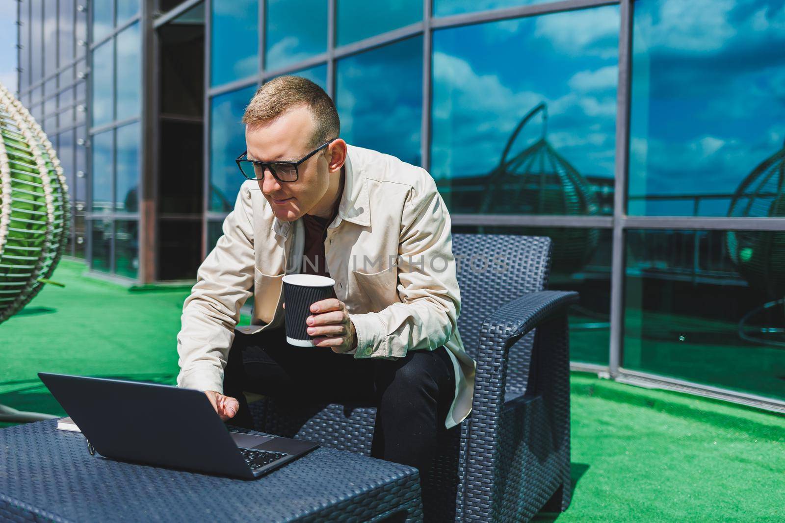 Confident young businessman, dressed in formal wear and stylish glasses, calls on a smartphone while sitting with coffee, on the terrace of a modern office building with a glass wall