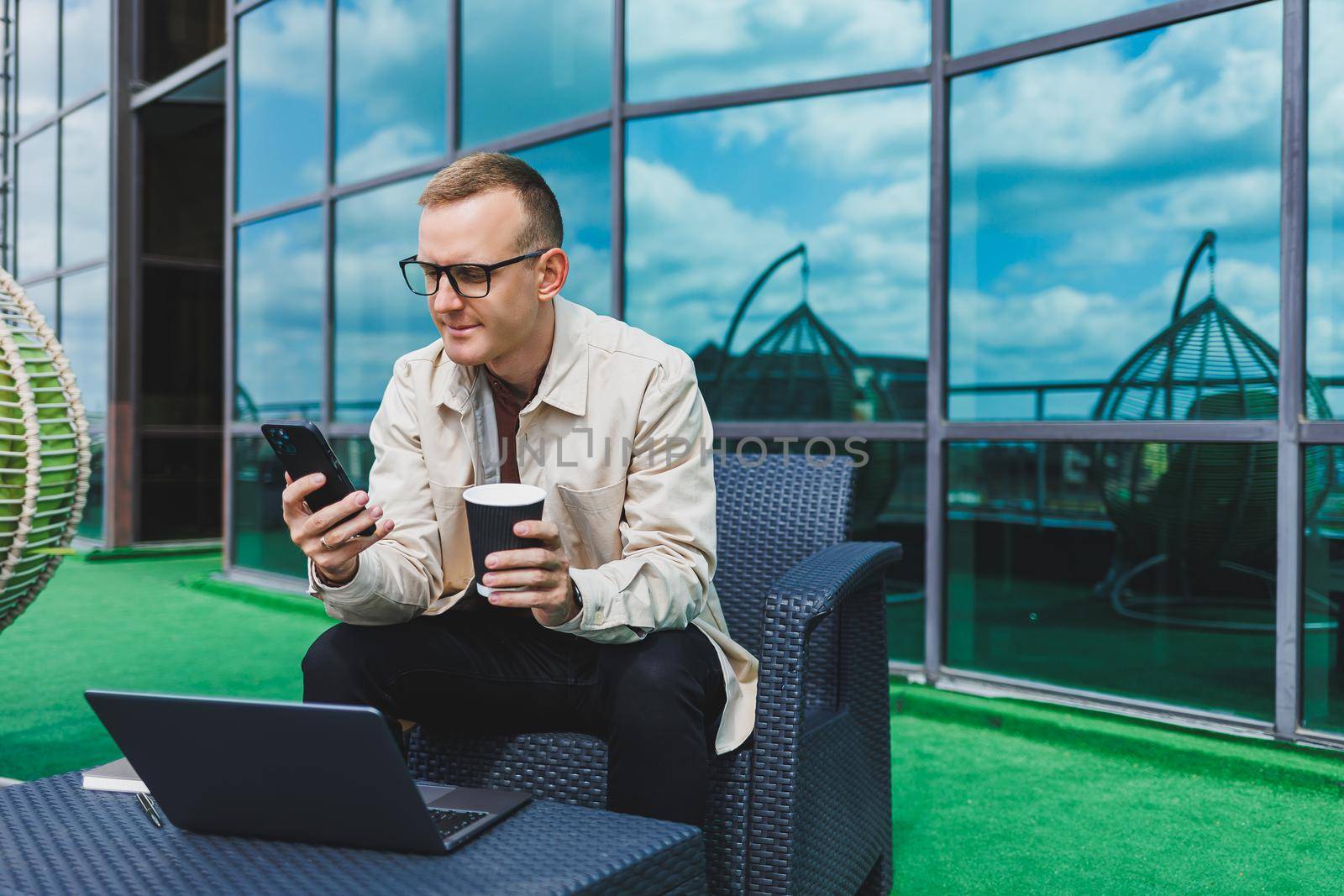 Confident young businessman, dressed in formal wear and stylish glasses, calls on a smartphone while sitting with coffee, on the terrace of a modern office building with a glass wall