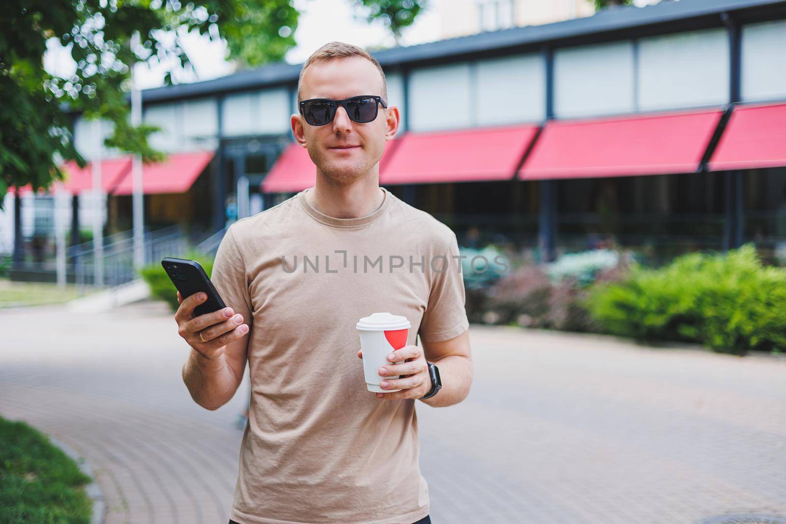 A bearded, smiling, stylish man in white shirt and sunglasses standing on the streets of the city and calling on mobile phone.