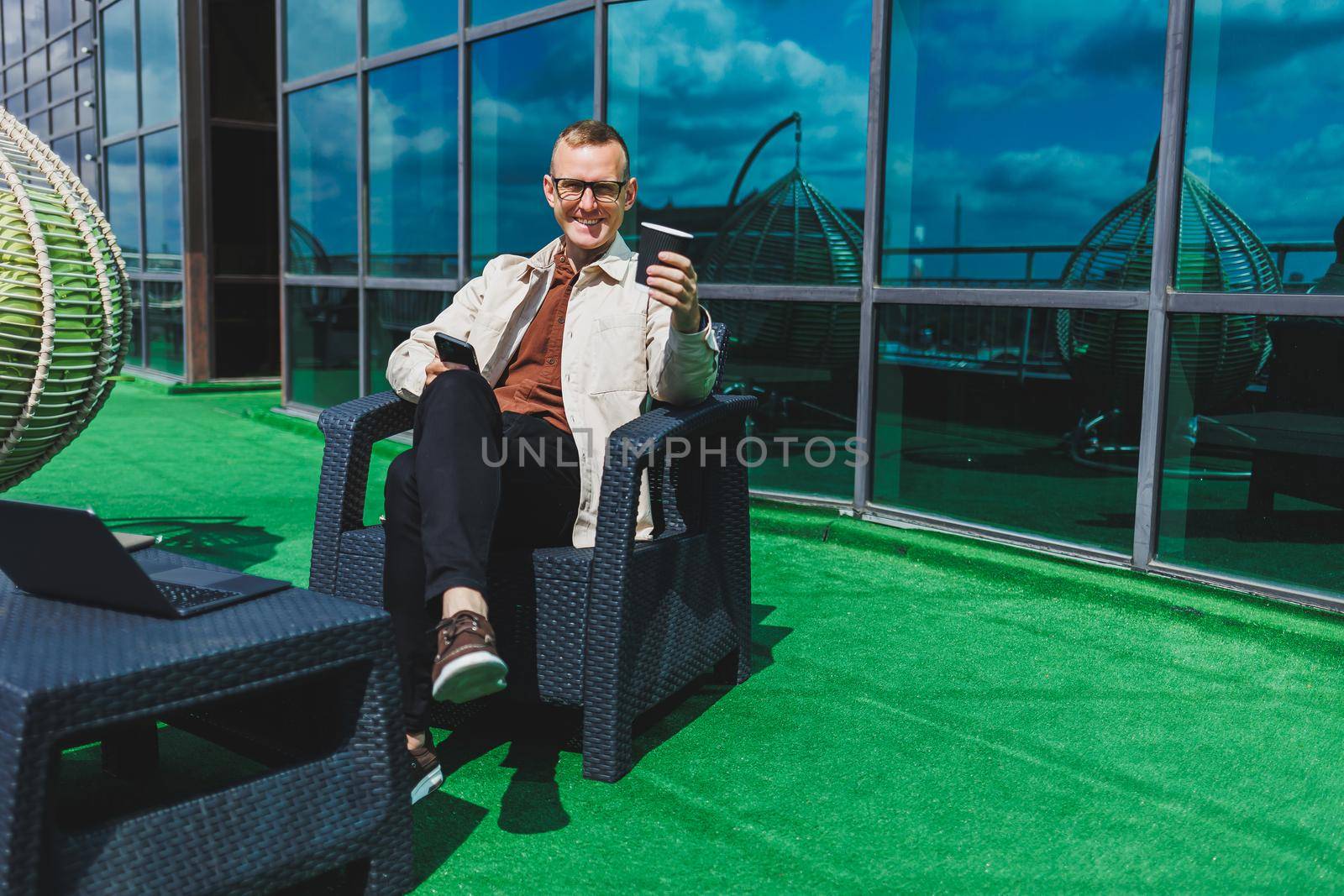 Confident young businessman, dressed in formal wear and stylish glasses, calls on a smartphone while sitting with coffee, on the terrace of a modern office building with a glass wall