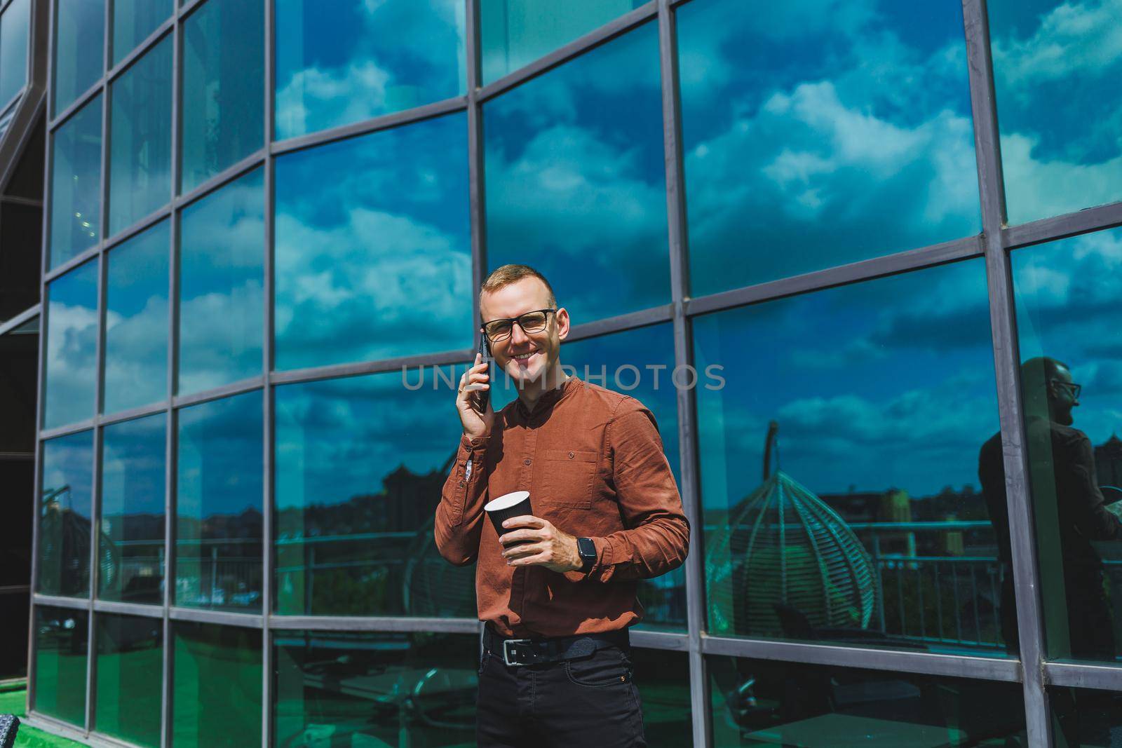 Attractive fashionable male businessman in a stylish shirt and trousers talking on the phone near the office building and drinking coffee, modern glass office