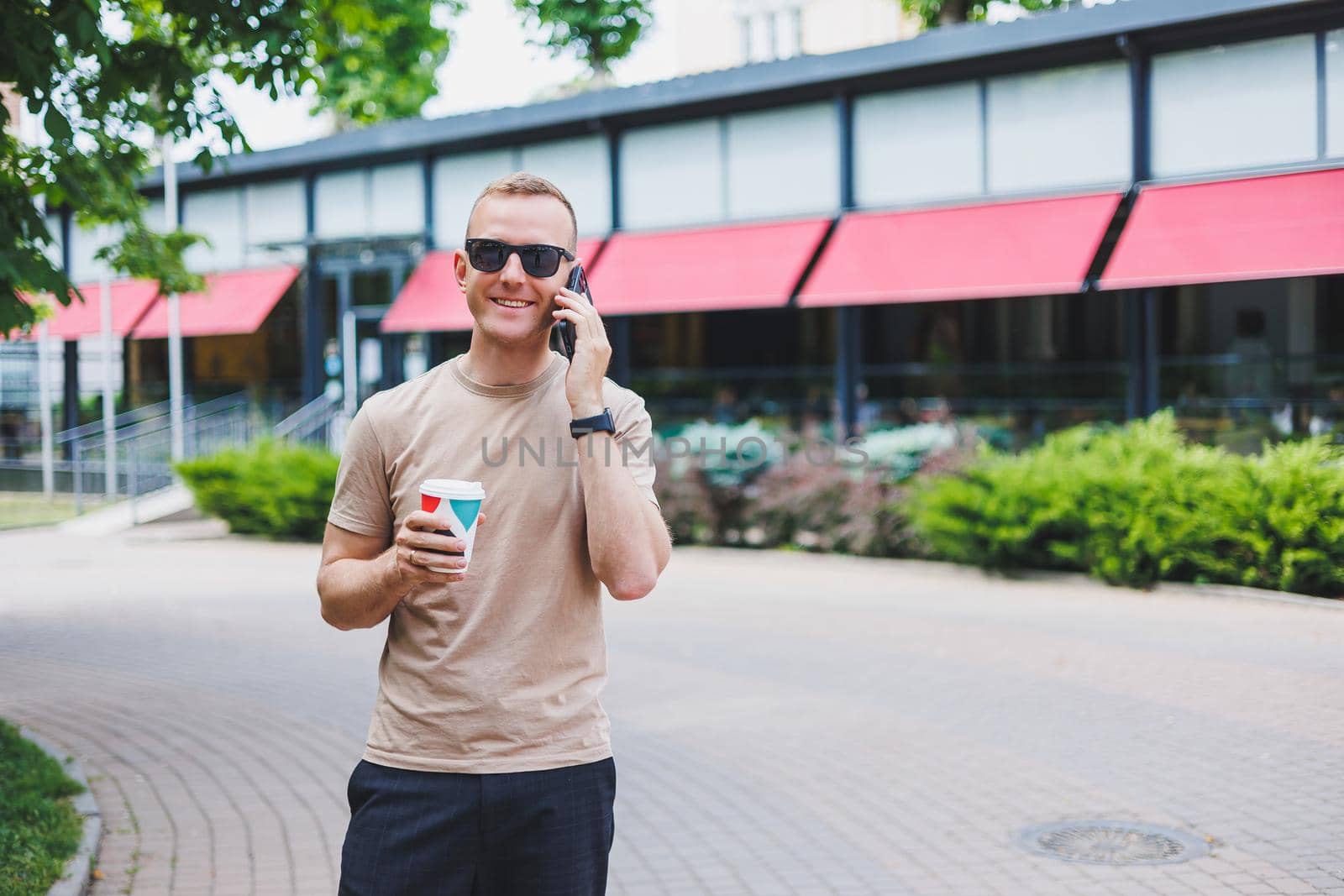 A bearded, smiling, stylish man in white shirt and sunglasses standing on the streets of the city and calling on mobile phone.