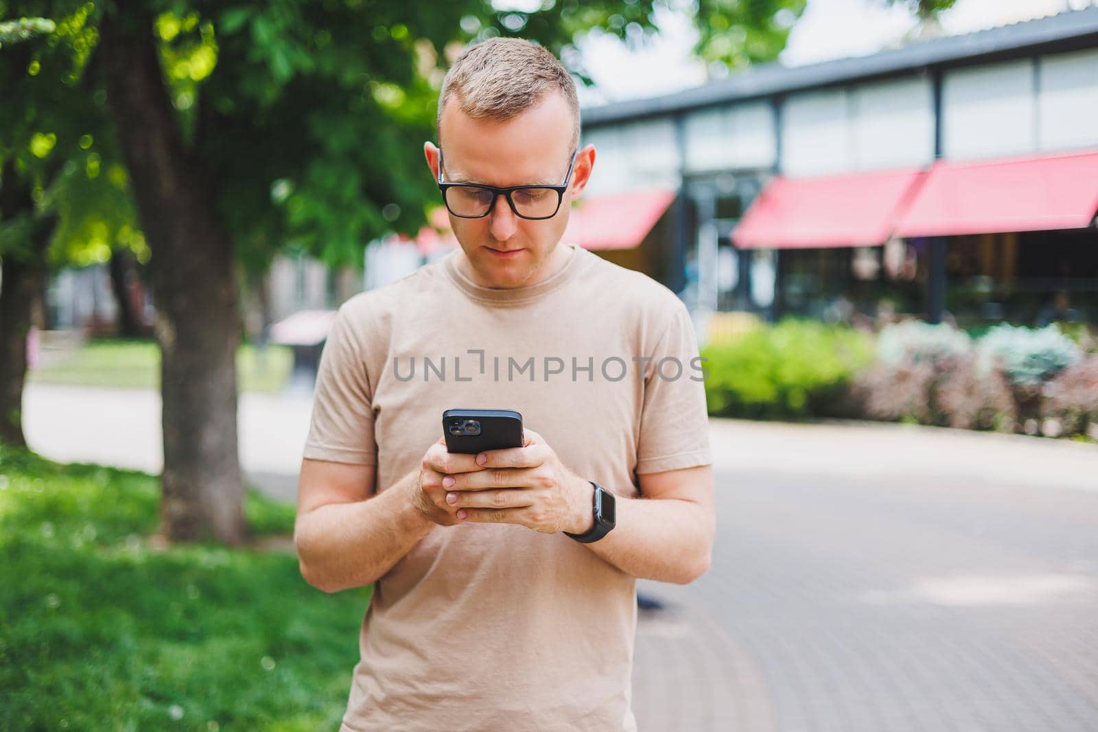 Portrait of handsome mature businessman holding messenger bag, wearing stylish eyeglasses looking at camera. Smiling middle aged man using smartphone, standing on the street. Mobile banking concept