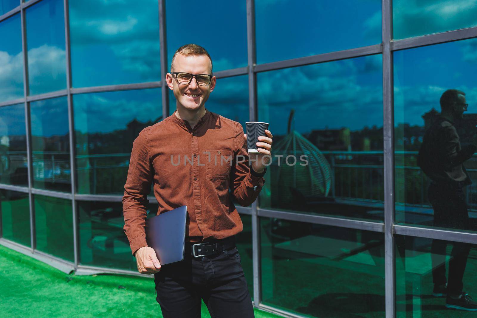 A young handsome attentive man in glasses with a laptop and a glass of coffee to go, stands on the terrace of an office. young smart developer