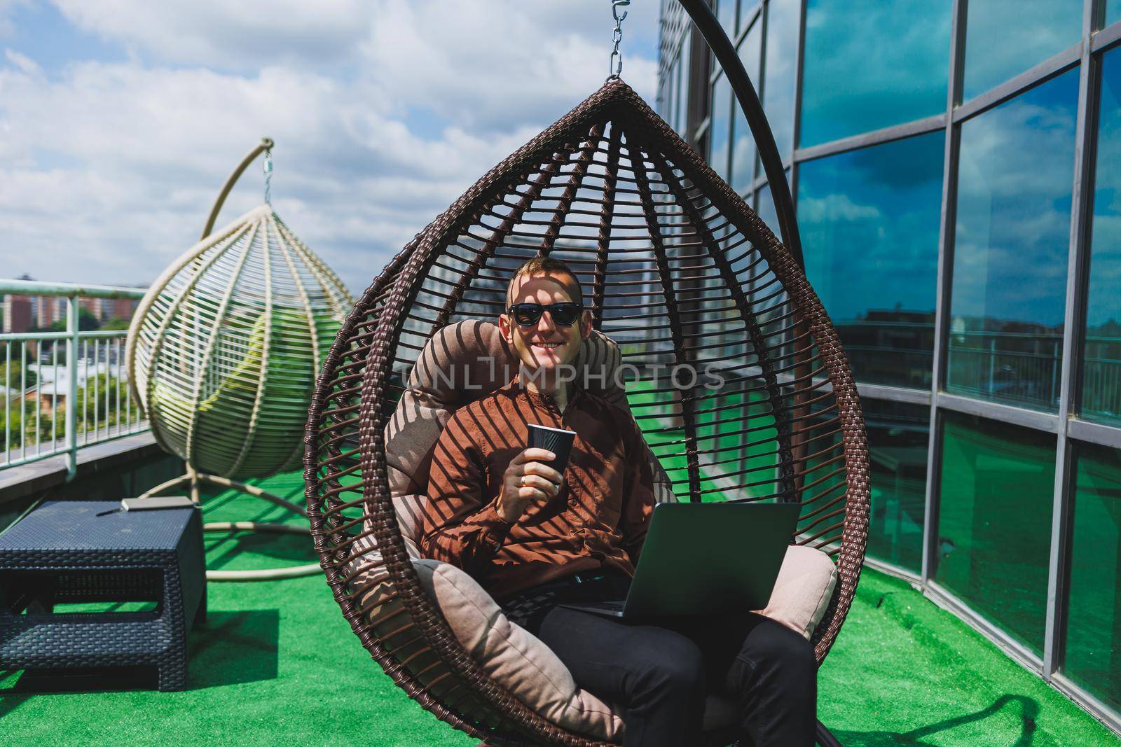 A businessman in formal wear sits on an armchair and looks at a laptop connected to a 4g wireless network on the terrace of a modern office while working remotely
