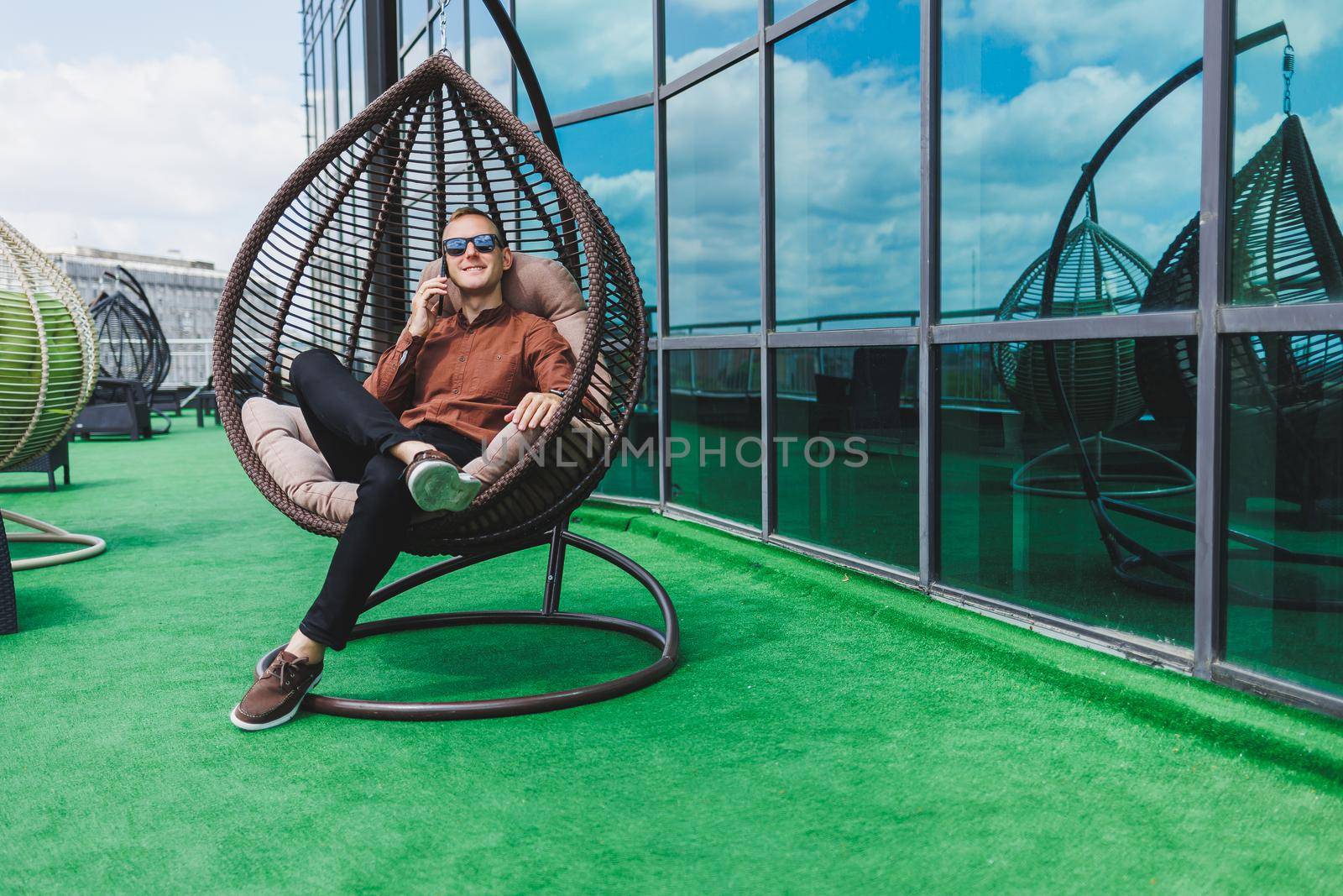 A cheerful young man in a shirt with a phone sits on the terrace of an office building after work, uses wireless internet, a positive male employee in glasses is talking on the phone
