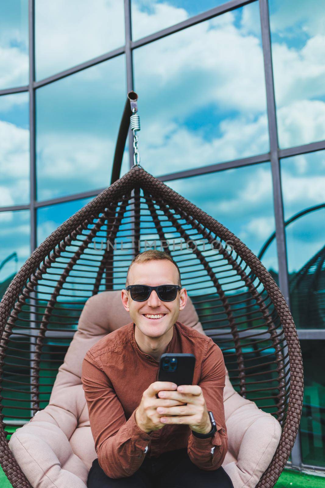A cheerful young man in a shirt with a phone sits on the terrace of an office building after work, uses wireless internet, a positive male employee in glasses is talking on the phone