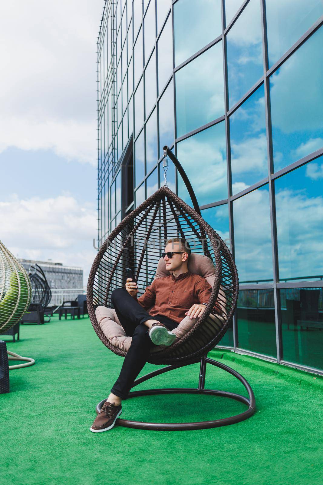 A cheerful young man in a shirt with a phone sits on the terrace of an office building after work, uses wireless internet, a positive male employee in glasses is talking on the phone