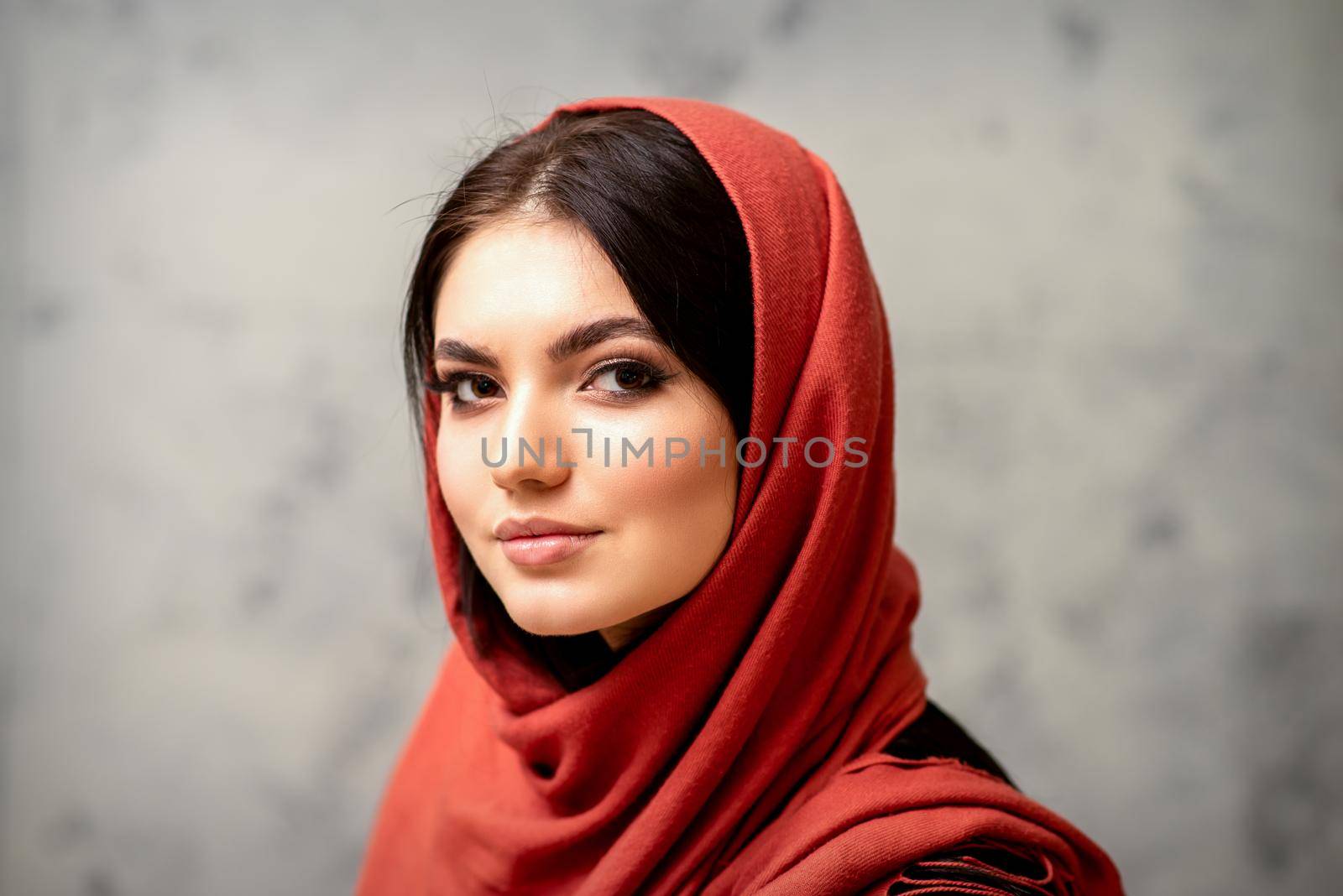 The fashionable young woman. Portrait of the beautiful female model with long hair and makeup in a red scarf. Beauty young caucasian woman on the background of a gray wall