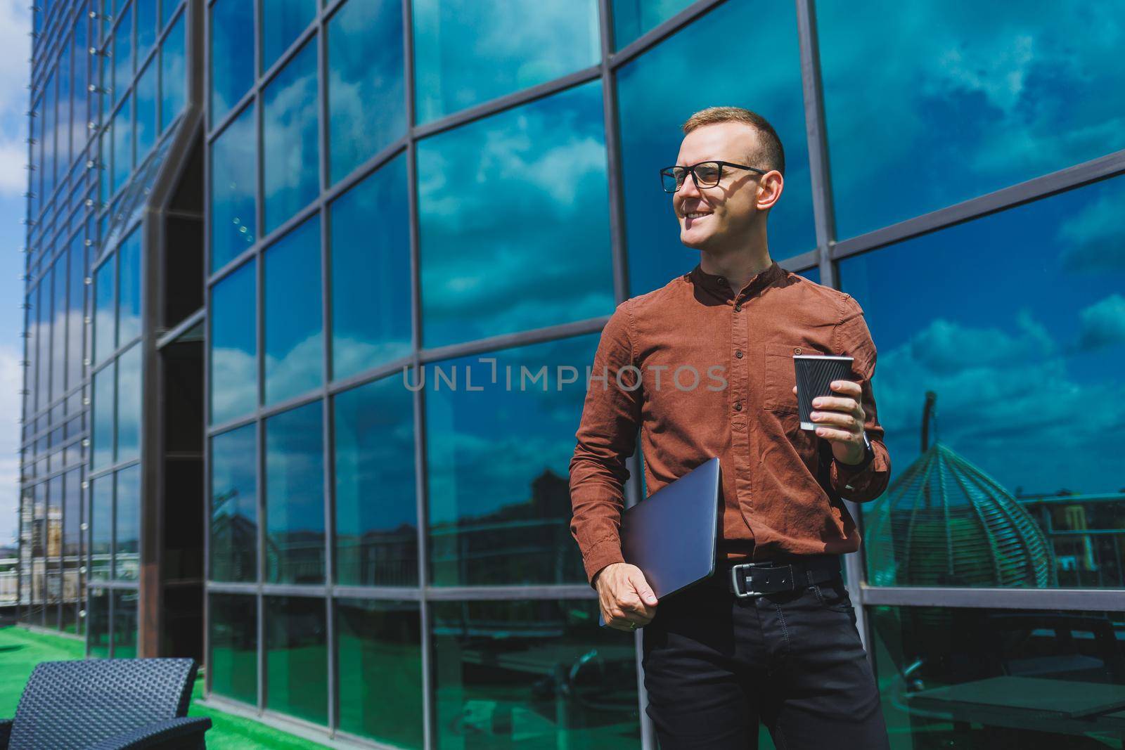 A young handsome attentive man in glasses with a laptop and a glass of coffee to go, stands on the terrace of an office. young smart developer