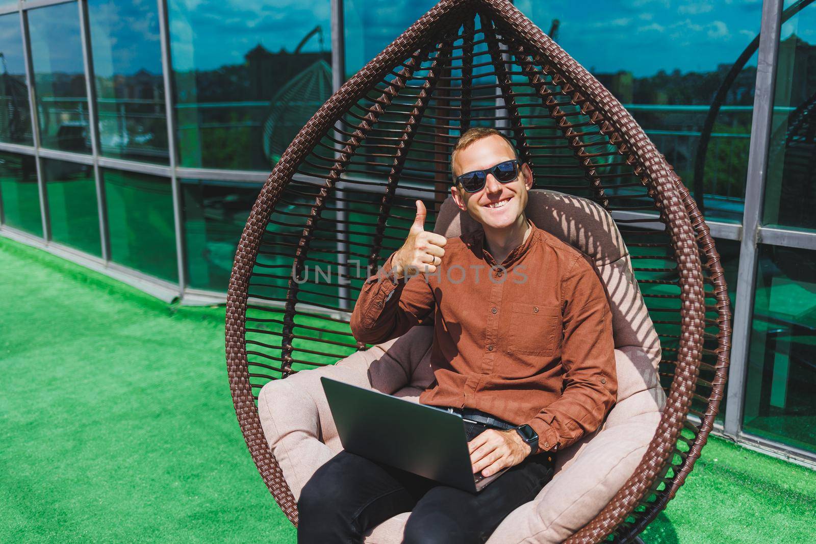 A businessman in formal wear sits on an armchair and looks at a laptop connected to a 4g wireless network on the terrace of a modern office while working remotely