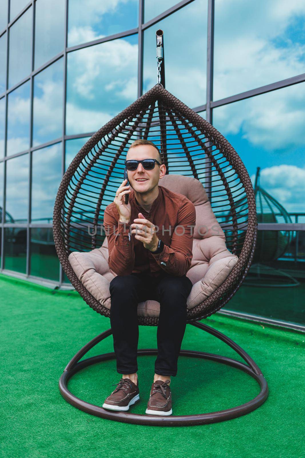 A cheerful young man in a shirt with a phone sits on the terrace of an office building after work, uses wireless internet, a positive male employee in glasses is talking on the phone